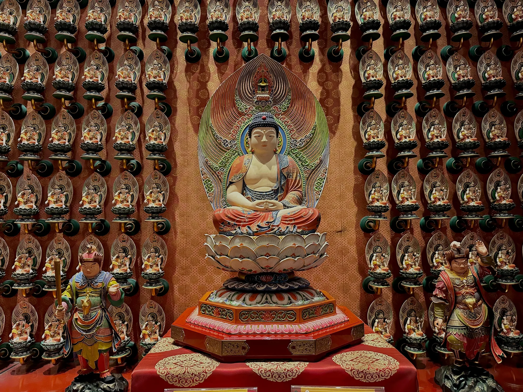 写真は、シンガポールで撮影された仏教寺院内部のものと思われる。中央には、蓮の台座に座った仏像が配置されている。仏像は、穏やかな表情で瞑想している。背景には、壁一面に等間隔で配置された多数の小さな仏像の装飾が見られる。これらの小さな仏像は、それぞれが独自の台座に取り付けられ、壁に固定されているようだ。壁自体は、伝統的な模様が施されたオレンジ色の壁紙で覆われている。写真の左端と右端には、護法神のような像が配置され、寺院の神聖な雰囲気を高めている。全体的に、写真は色彩豊かで、細部にまでこだわった装飾が印象的である。寺院の荘厳さと静けさを伝えている。
