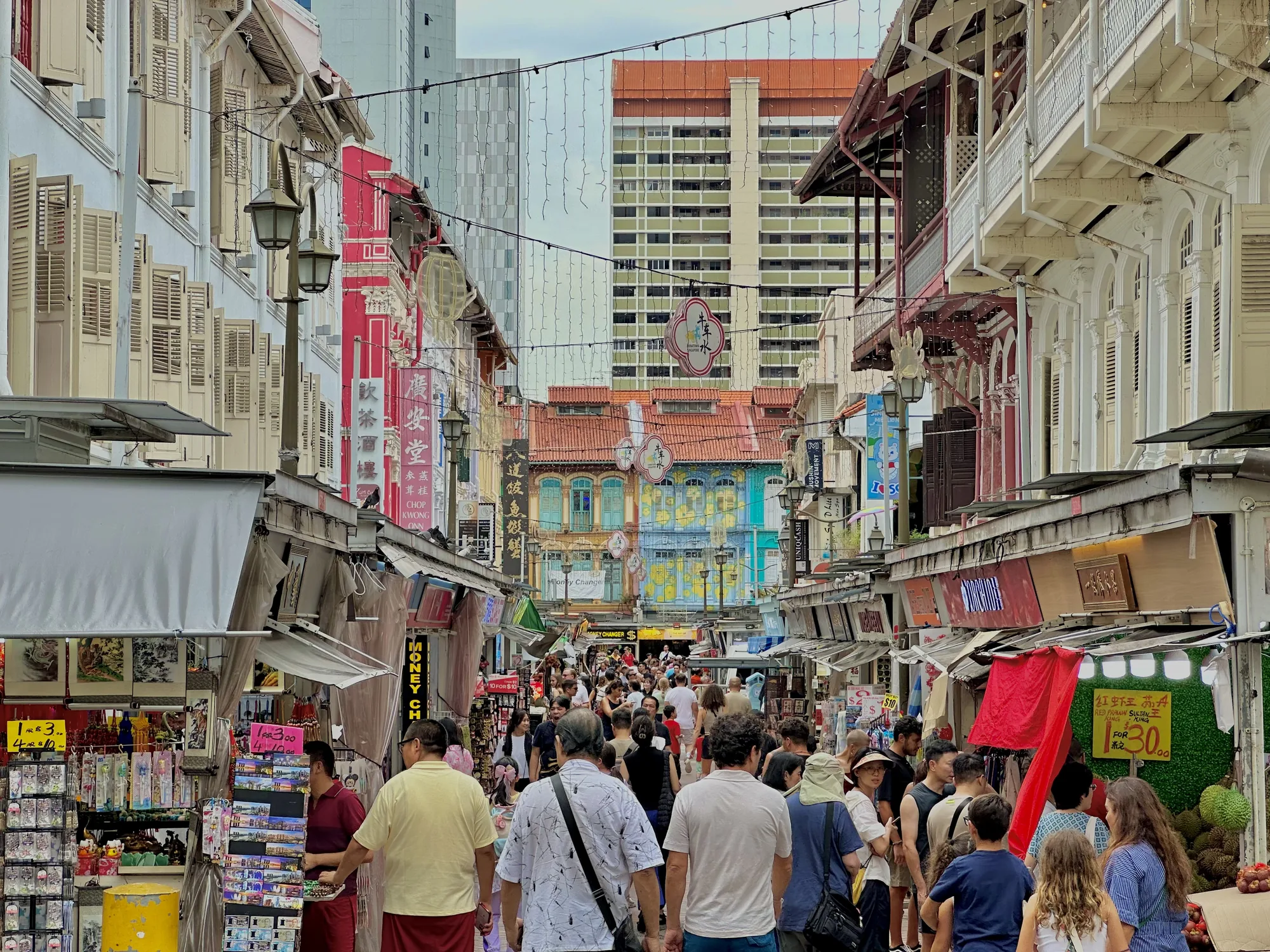 写真はシンガポールの不明な場所で撮影された、賑やかな通りを捉えています。通りには多くの人々が行き交い、両側には様々な店舗や屋台が並んでいます。建物の外観はカラフルで、歴史を感じさせるデザインが特徴的です。特に、鮮やかな赤色や青色の建物が目を引きます。屋台では様々な商品が販売されており、食品や雑貨などが見られます。通り全体は活気にあふれ、観光客や地元の人々で賑わっている様子が伝わってきます。