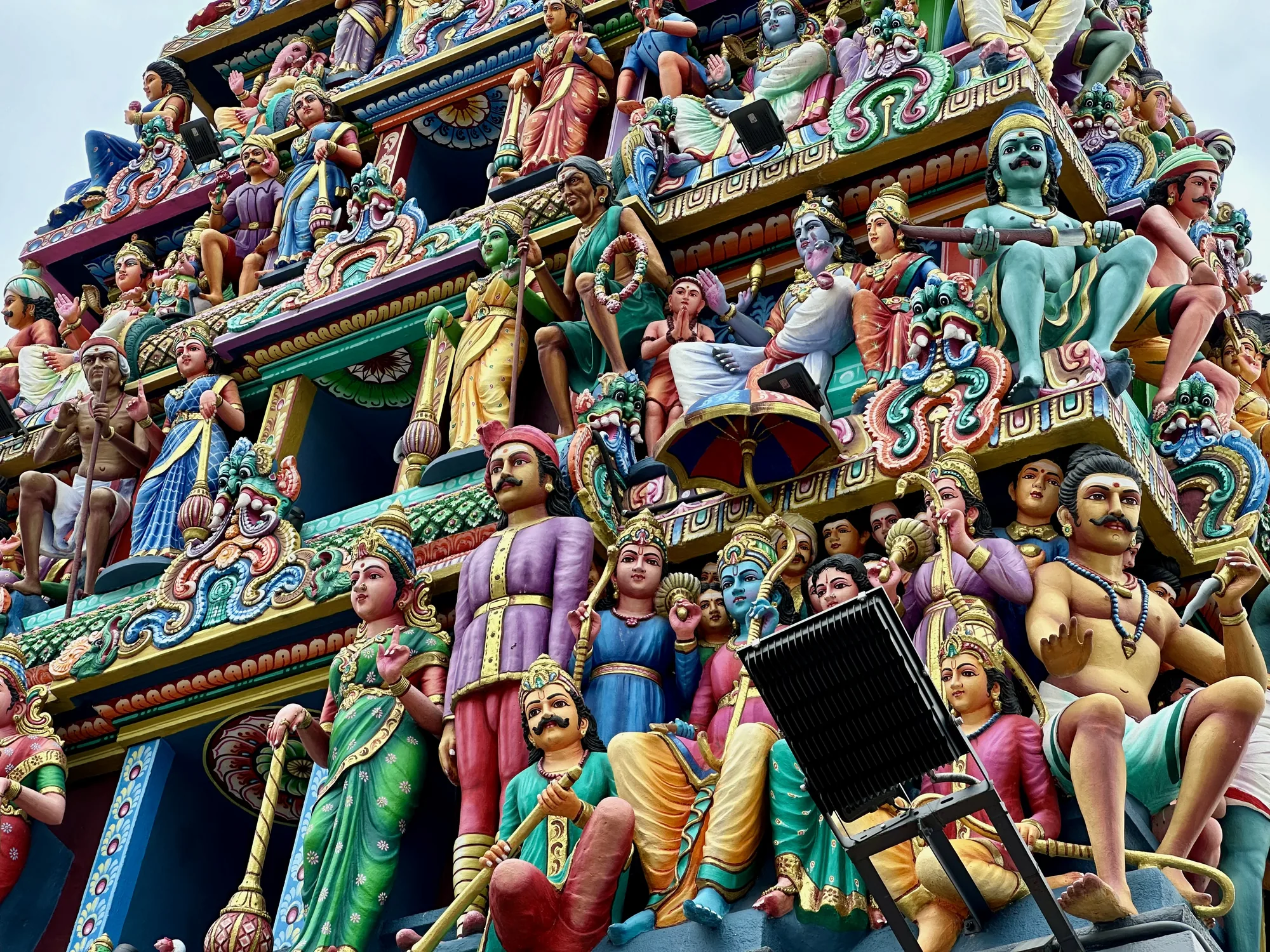 写真は、シンガポールのヒンドゥー寺院の鮮やかな彫刻をクローズアップで捉えています。寺院の屋根部分に、神々や人物を象った色とりどりの像が密集して配置されており、青、緑、赤、黄など、鮮やかな色彩が目を引きます。それぞれの像は細部まで丁寧に作られており、服装や装飾品、表情など、豊かな表現力を持っています。層状に重なった彫刻群は、壮観で華やかな印象を与えます。また、写真の右下には、寺院の修復作業で使用されると思われる照明器具が写っており、文化財の維持活動の一端を垣間見ることができます。