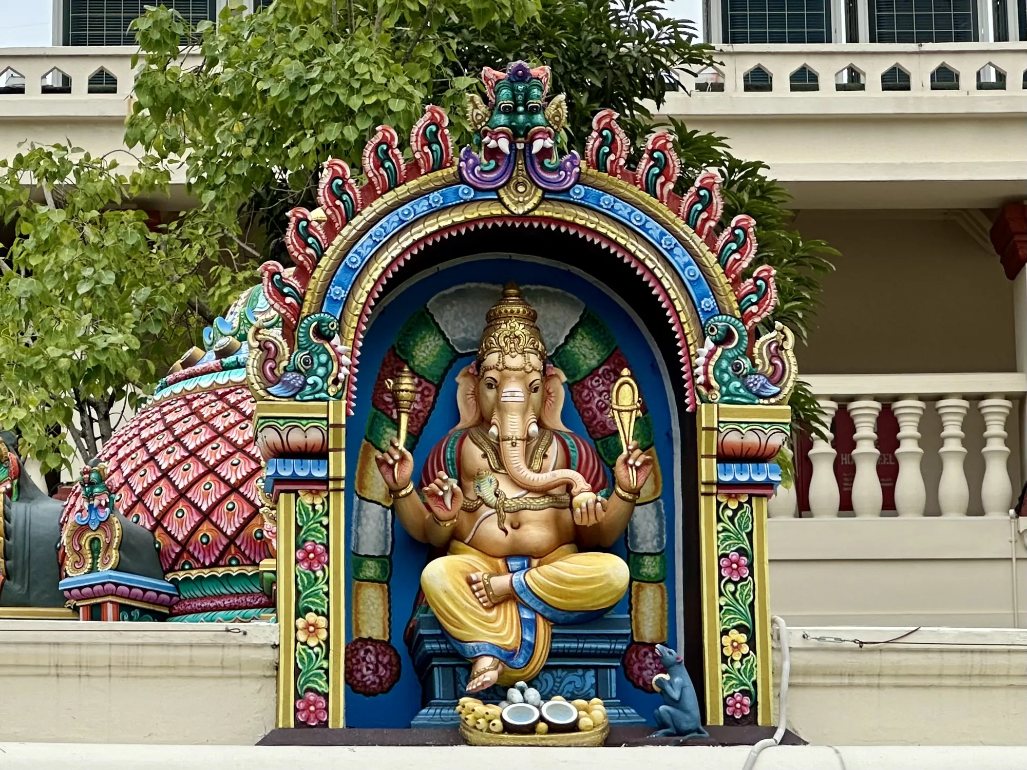 写真には、シンガポールのどこかで撮影されたと思われる、ヒンドゥー教の神であるガネーシャの像が写っています。像は鮮やかな色彩で装飾されており、精巧な彫刻が施されたアーチ型の構造の中に安置されています。ガネーシャは蓮華座に座り、手には神具を持っています。足元には、供え物と思われる果物とココナッツ、そしてガネーシャの乗り物であるネズミの像が置かれています。  背景には建物の一部と緑豊かな木々が見え、都会の中にある寺院や宗教施設の一角であることが示唆されます。全体的に、この写真はシンガポールの多文化性と宗教的な多様性を象徴しているように見えます。鮮やかな色彩と細部までこだわった彫刻は、見る人に強い印象を与え、異文化への関心を抱かせます。