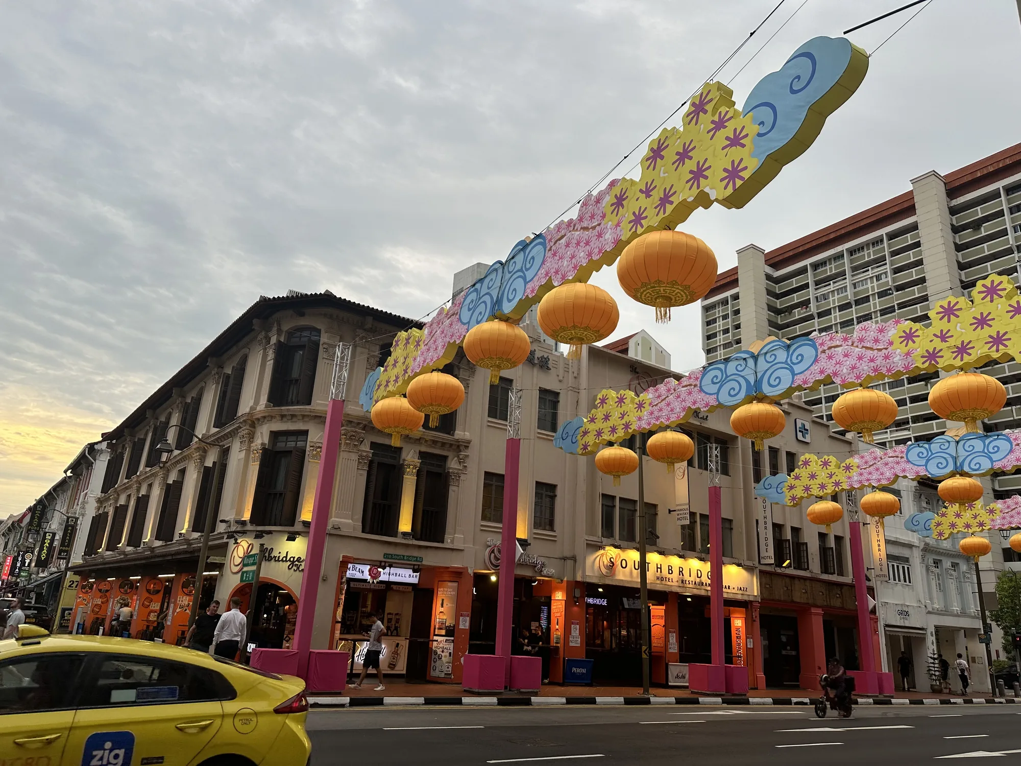 写真はシンガポールの街並みを捉えたものです。画面奥には歴史を感じさせる白い建物が並び、その上にグレーがかった空が広がっています。建物の間には、祭りの飾り付けと思われる、雲の形をした青色の装飾や、黄色とピンクの花柄の装飾が施されています。また、オレンジ色の提灯が通りに沿って吊るされ、華やかな雰囲気を醸し出しています。道路には黄色いタクシーが走っており、歩道には人々が歩いています。手前の建物にはレストランやバーなどの店があり、夜に向けて賑わいを見せているようです。全体的に、古き良きシンガポールの街並みと現代的な要素が混ざり合った、魅力的な光景が写し出されています。