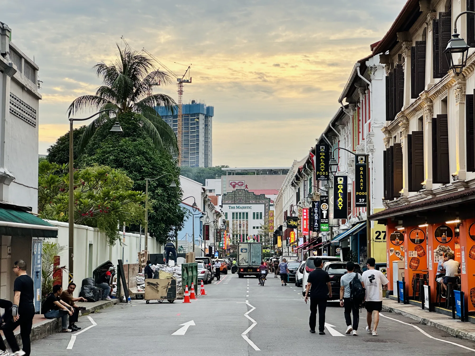写真はシンガポールの街並みを捉えたものです。視線は奥へと伸びる道路に沿って誘導され、道路には人々が歩き、車が走行しています。道路の両側にはカラフルな建物が立ち並び、右側には伝統的なショップハウスが並んでいます。これらの建物は、独特な装飾が施された窓枠やルーバー、柱などが特徴的です。左側にはもう少しシンプルな建物が見え、緑豊かな木々が並んでいます。道路には白い矢印が描かれており、交通の方向を示しています。道路脇には工事現場のような場所があり、オレンジ色のコーンが置かれています。遠くには建設中の高層ビルがあり、クレーンがそびえ立っています。空は曇り空で、夕焼けのような淡い光が射し込んでいます。全体的に、活気がありながらも落ち着いた雰囲気の写真です。OCRの結果から、「The Majestic」、「Galaxy Pods 30A Mosque St」、「Victoria」といった看板が確認できます。また、写真の場所は「27 Mosque Street」付近であることがわかります。