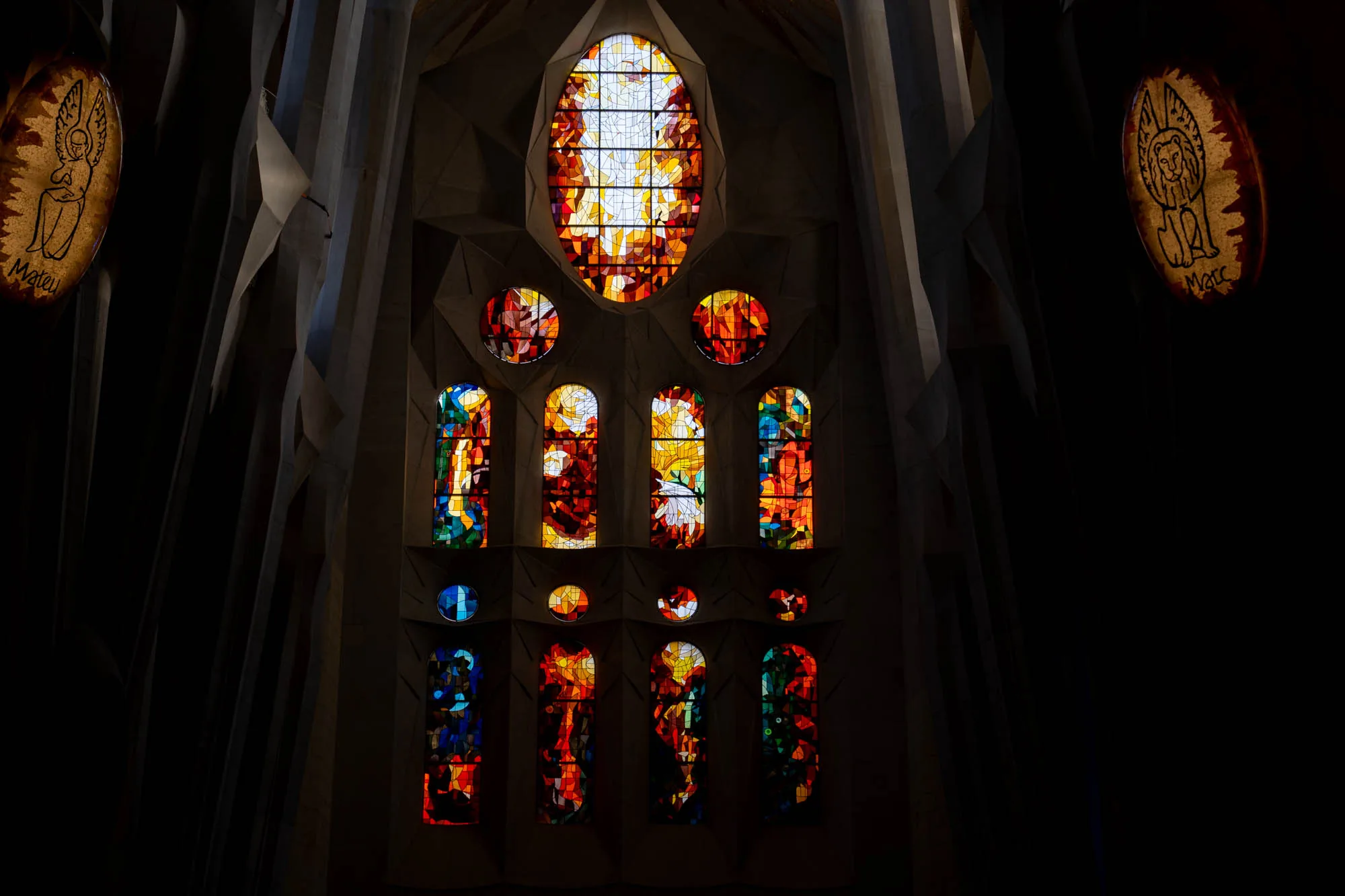 スペイン・バルセロナにあるサグラダファミリア内部を撮影した写真です。写真の中心には、複数のステンドグラスが配置されています。上部には大きな楕円形のステンドグラスがあり、白とオレンジ色の光が印象的です。その下には、円形や縦長のステンドグラスが並び、赤、オレンジ、黄色、青など様々な色が使われています。ステンドグラスを通して差し込む光が、教会内部に幻想的な雰囲気を作り出しています。画面の左右にはそれぞれ翼の生えた人物（おそらく天使）の絵が描かれた楕円形の飾りが配置されており、「Mateu」「Marc」という文字が記されています。全体的に暗めの照明で、ステンドグラスの色彩が際立って見える構図です。