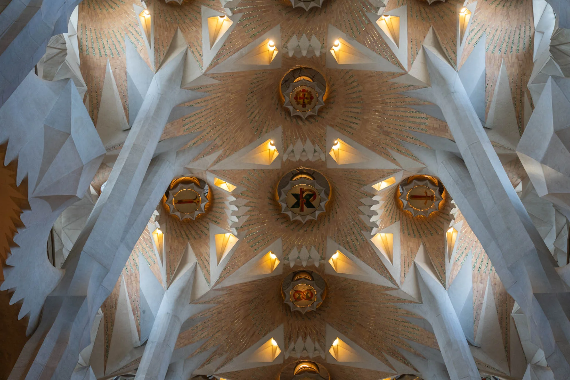 写真は、スペインのバルセロナにあるサグラダファミリアの天井を捉えたものです。天井は幾何学的なデザインで、複雑な模様が施されています。淡いベージュ色と白色が基調で、光沢のある装飾が施され、中心には紋章のような装飾が埋め込まれています。複数のスポットライトが設置され、天井全体を照らし出しています。建築様式は非常に独創的で、アントニ・ガウディの設計思想が色濃く反映されていることが伺えます。全体的に荘厳で神秘的な雰囲気を醸し出しています。