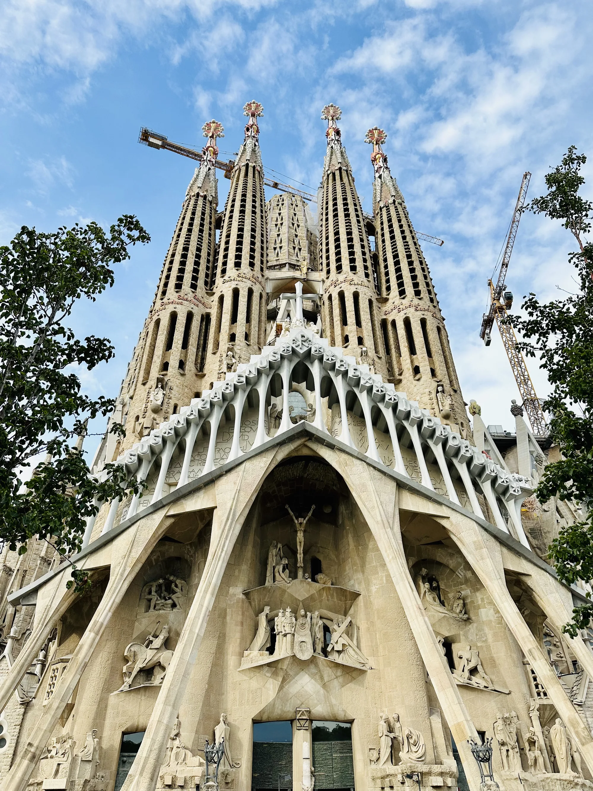 写真はスペインのバルセロナにあるサグラダファミリアの堂々たる外観を捉えています。下から見上げるように撮影された構図で、建物の圧倒的な高さと細部にわたる彫刻が強調されています。空は青く、ところどころに白い雲が浮かんでおり、建物の輪郭を際立たせています。建物の石の色は明るいベージュで、長年の風雨に晒された様子がうかがえます。  サグラダファミリアの尖塔は高くそびえ立ち、それぞれに複雑な装飾が施されています。尖塔の上部には、特徴的な装飾が施されており、建設に使われているクレーンがいくつか見えます。建物の正面には、キリストの生涯や聖書の物語を描いた彫刻が数多く施されており、その芸術性の高さに目を見張ります。建物の下部には、アーチ状の構造や彫刻が施された壁面があり、ゴシック様式とモデルニスモ様式が融合した独特のデザインが印象的です。  写真全体から、サグラダファミリアの壮大さ、建設の途方もない規模、そしてガウディの独創的なビジョンが伝わってきます。建設中のクレーンは、この壮大なプロジェクトが現在も進行中であることを示唆しており、完成への期待を高めます。