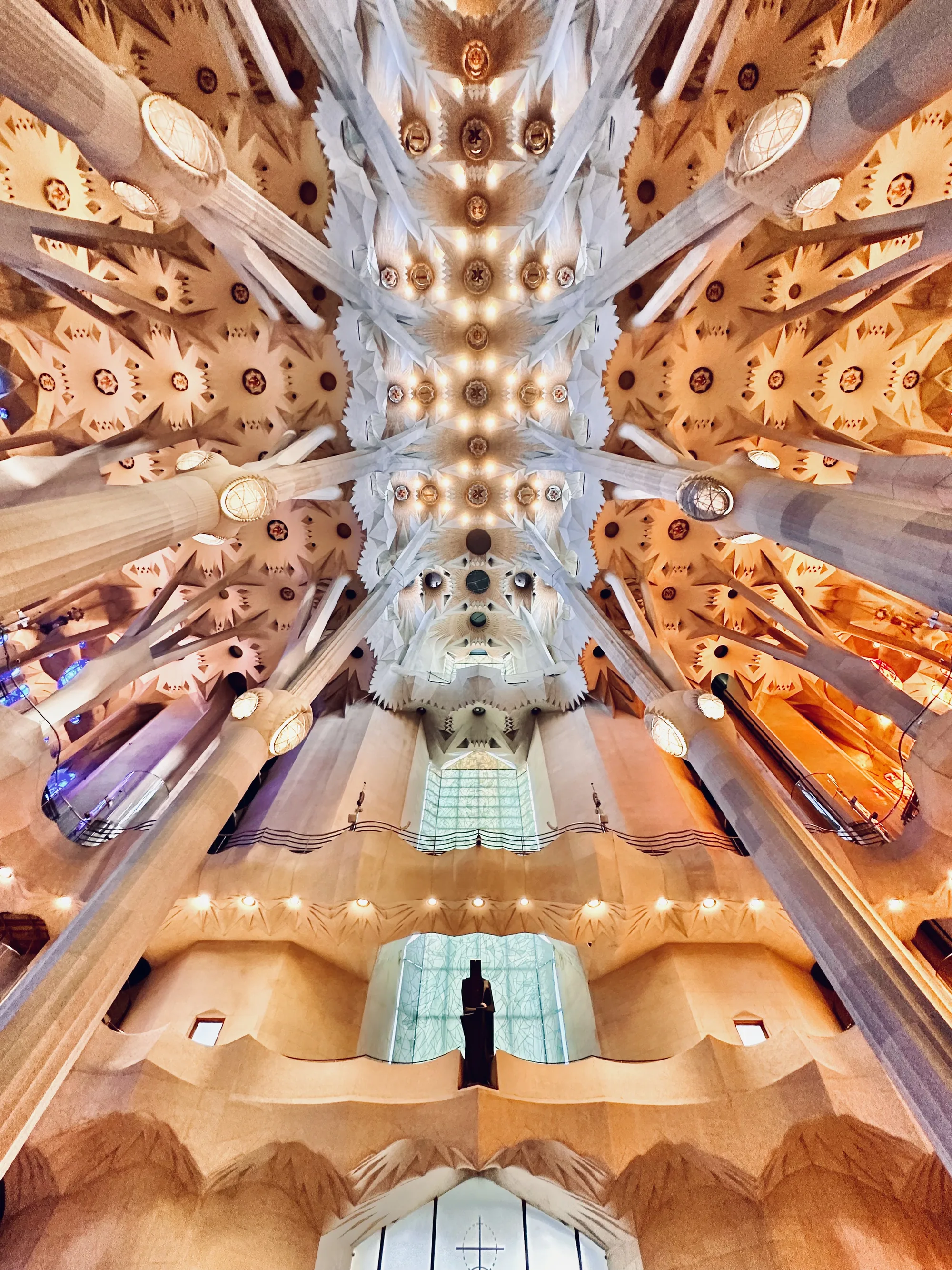 スペイン・バルセロナのサグラダファミリア内部を撮影した写真です。写真の視点は低い位置から天井を見上げており、高い天井を支える柱が何本もそびえ立ち、その複雑な構造が印象的です。柱は上に向かって枝分かれしており、天井は星のような幾何学的な模様で埋め尽くされています。全体的にベージュや白を基調とした色使いで、照明が暖かく照らしており、神聖で荘厳な雰囲気を醸し出しています。写真の中央奥には窓があり、その前に人影のような像が立っています。独特な視点と構造美が組み合わさり、サグラダファミリアの壮大さを感じさせる一枚です。