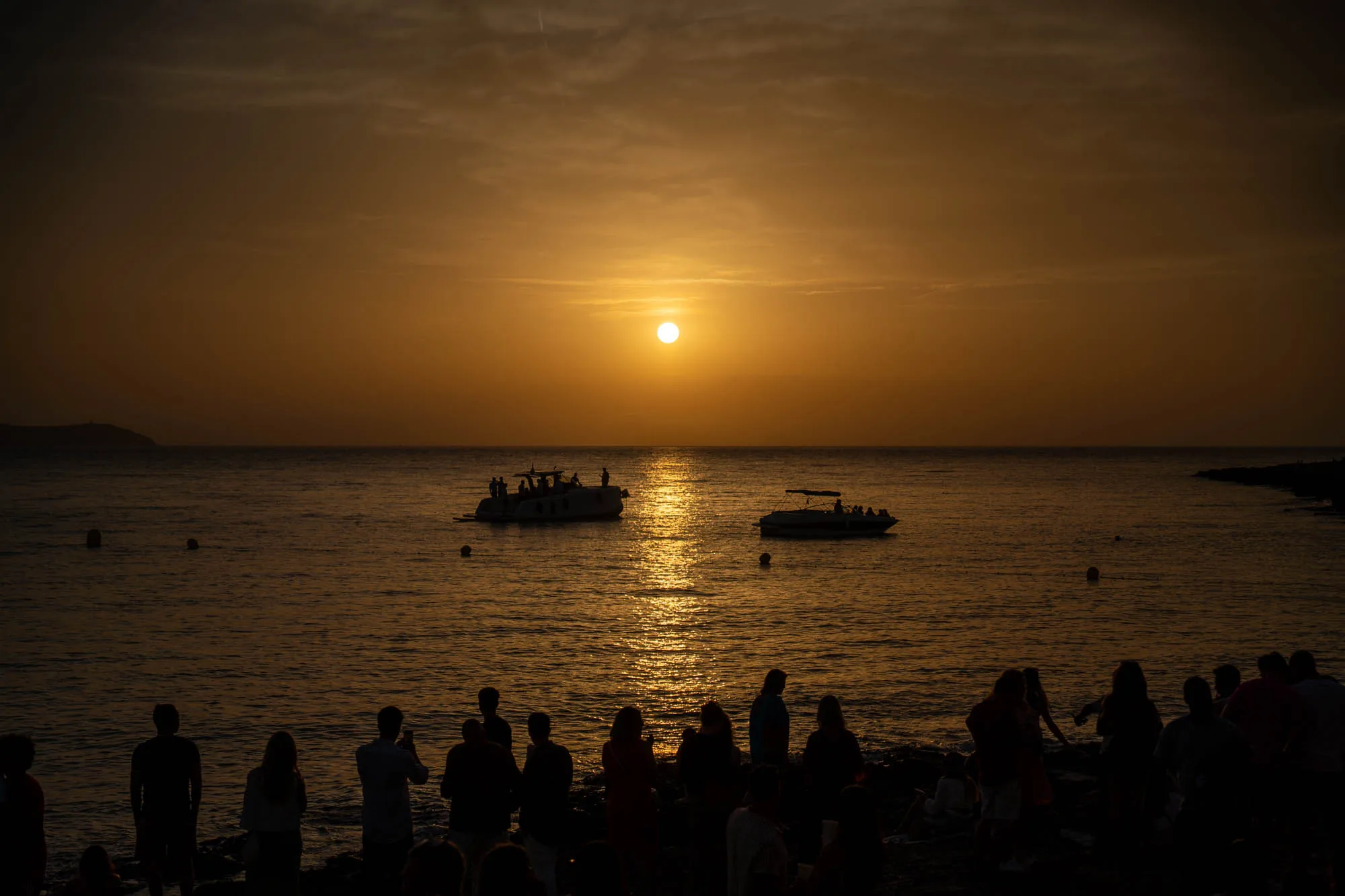 写真はスペインのイビサ島で撮影された夕焼けの風景です。空全体がオレンジ色に染まり、太陽が水平線近くに沈んでいます。海面は夕日の光を反射して金色に輝き、きらめいています。海には2隻のボートが浮かんでおり、それぞれに人が乗っている様子がシルエットで見えます。手前の海岸には、夕日を眺める人々のシルエットが多数写っています。人々は海岸に集まり、夕焼けの美しい景色を写真に収めたり、楽しんだりしているようです。全体的に、静かで穏やかな雰囲気の、美しい夕暮れの瞬間を捉えた写真です。