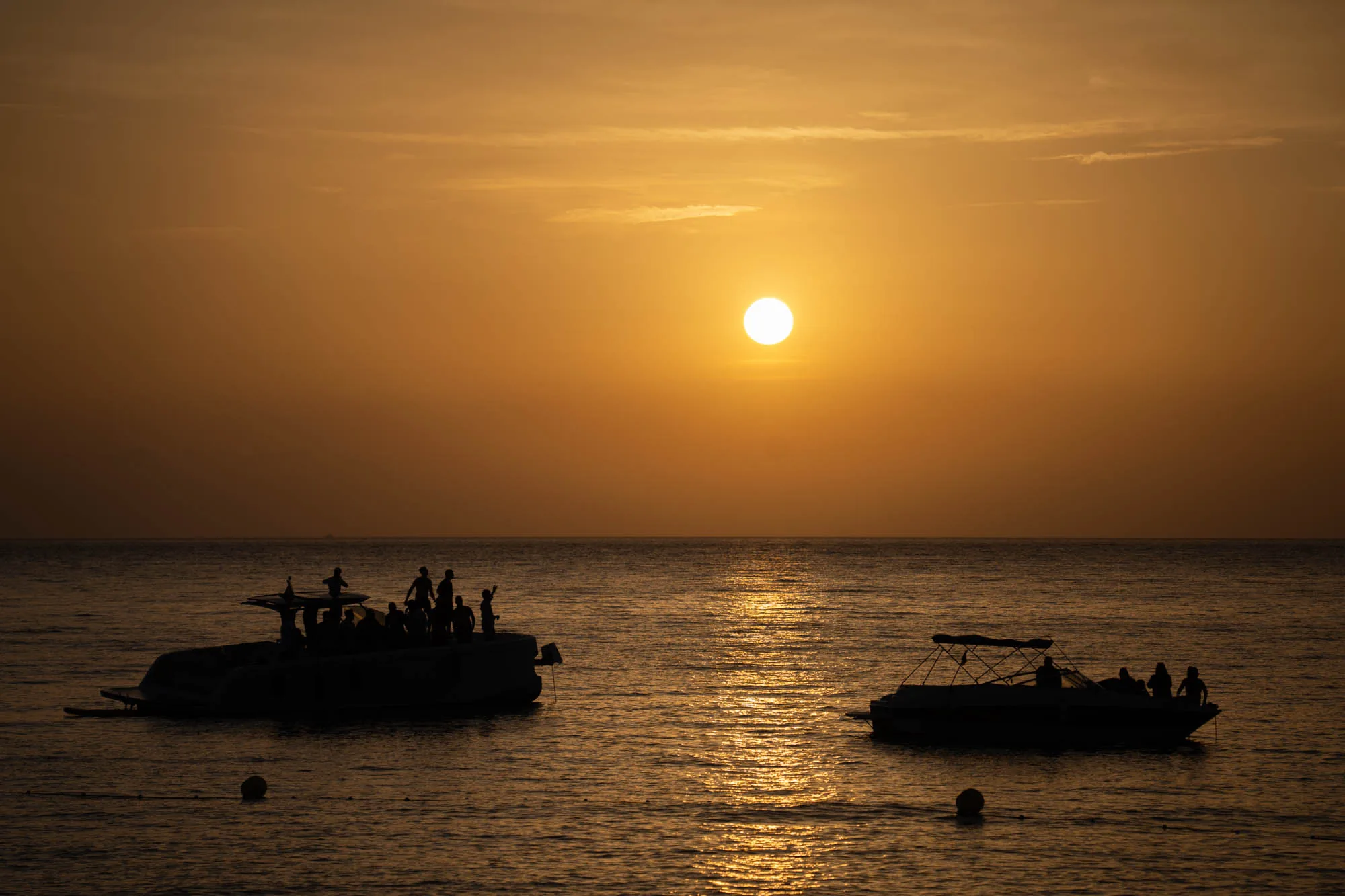 この写真はスペインのイビサ島で撮影された夕焼けの海の風景です。画面全体が夕日の暖かく優しいオレンジ色に染まっており、水平線に沈む太陽が輝いています。海面は太陽の光を反射してキラキラと輝き、穏やかな波が打ち寄せています。画面左側と右側にはそれぞれボートが浮かんでいます。どちらのボートにも複数の人影があり、夕日を眺めたり、ポーズをとったりしているようです。ボートのシルエットが夕焼けの空に浮かび上がり、幻想的な雰囲気を醸し出しています。全体的に穏やかで美しい、リラックスした時間が流れているような印象を与える写真です。