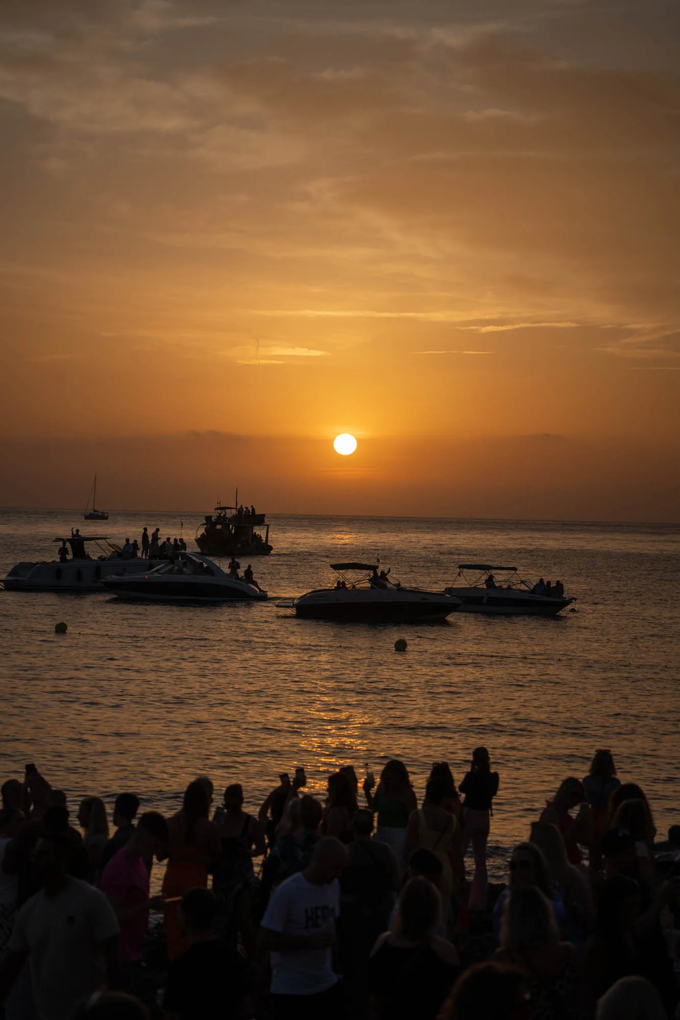 この写真はスペイン、イビサ島の夕暮れ時を捉えたものです。空はオレンジ色に染まり、太陽が水平線に沈みかけています。海面も夕日に照らされ、金色に輝いています。海には複数のボートが浮かんでおり、そのうちのいくつかには人が乗っているのが見えます。手前の海岸には大勢の人々が集まっており、夕日を眺めたり、会話を楽しんだりしているようです。人々のシルエットが夕焼けに浮かび上がり、幻想的な雰囲気を醸し出しています。全体的に、穏やかでリラックスした、イビサの美しい夕暮れの風景が描写されています。