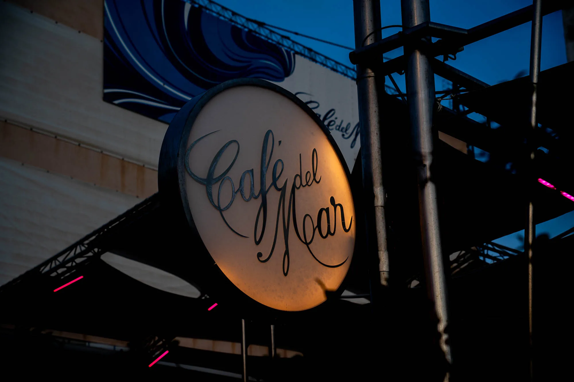 写真は、スペインのイビサ島にある有名なカフェ「Cafe del Mar」の看板を捉えています。夕暮れ時の空を背景に、丸い看板がオレンジ色の光を放ち、その上に手書き風の優雅なフォントで「Cafe del Mar」と書かれています。看板は金属製の骨組みに取り付けられており、背景には建物の構造物や他の看板の一部が見えます。全体的に暗い色調で、夕暮れ時の落ち着いた雰囲気と、カフェの洗練された雰囲気を醸し出しています。