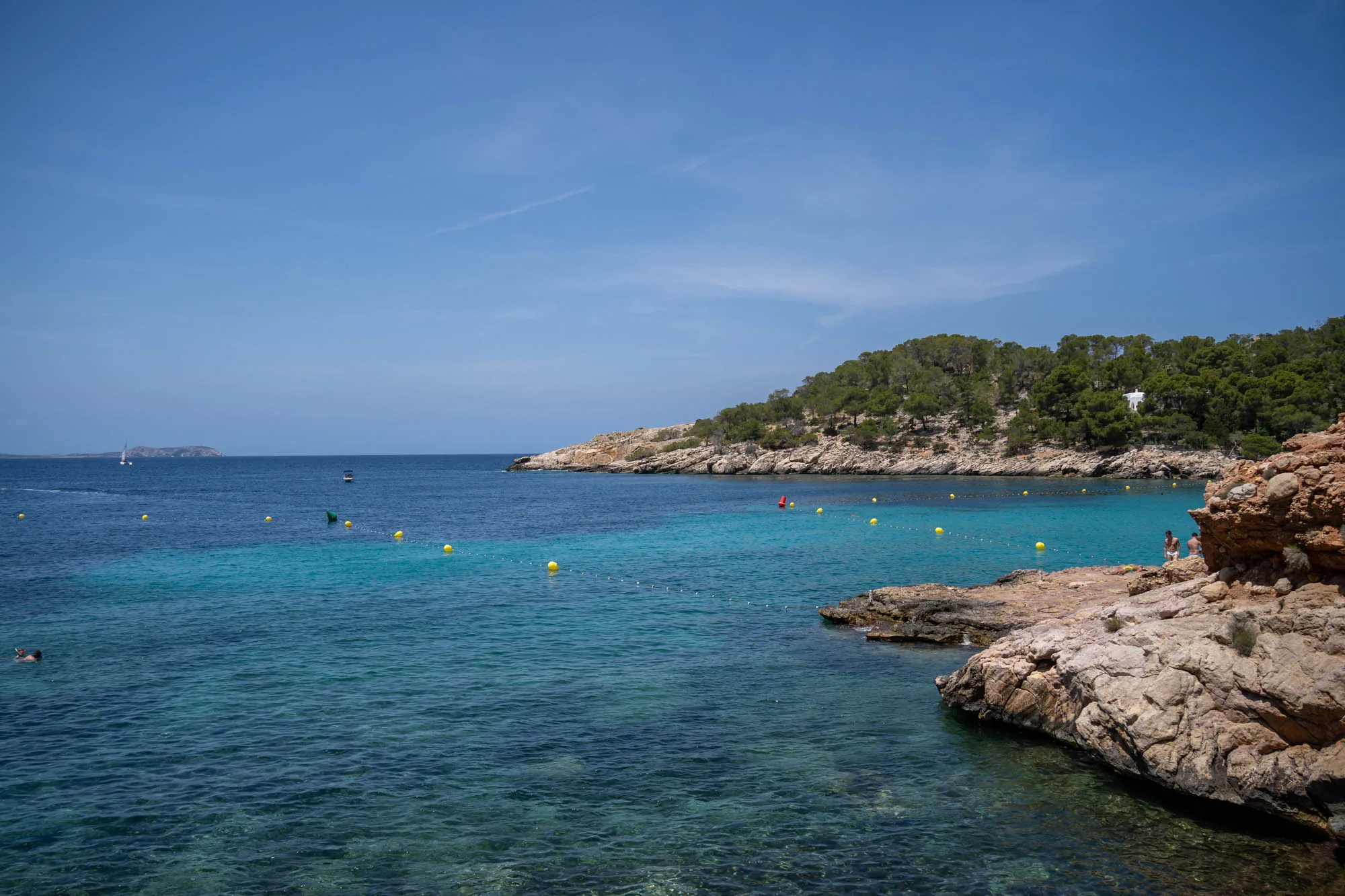写真はスペインのイビサ島で撮影された風景です。明るい青空の下、透明度の高いターコイズブルーの海が広がっています。海面には黄色いブイが浮かび、安全な遊泳区域を示しているようです。遠くには小さな島と白い帆のヨットが見えます。海岸線は岩場で、木々が生い茂る丘へと続いています。手前の岩場には人が座っている様子も見られます。全体的に、穏やかでリラックスした雰囲気の、イビサ島の美しいビーチの風景を捉えた写真です。