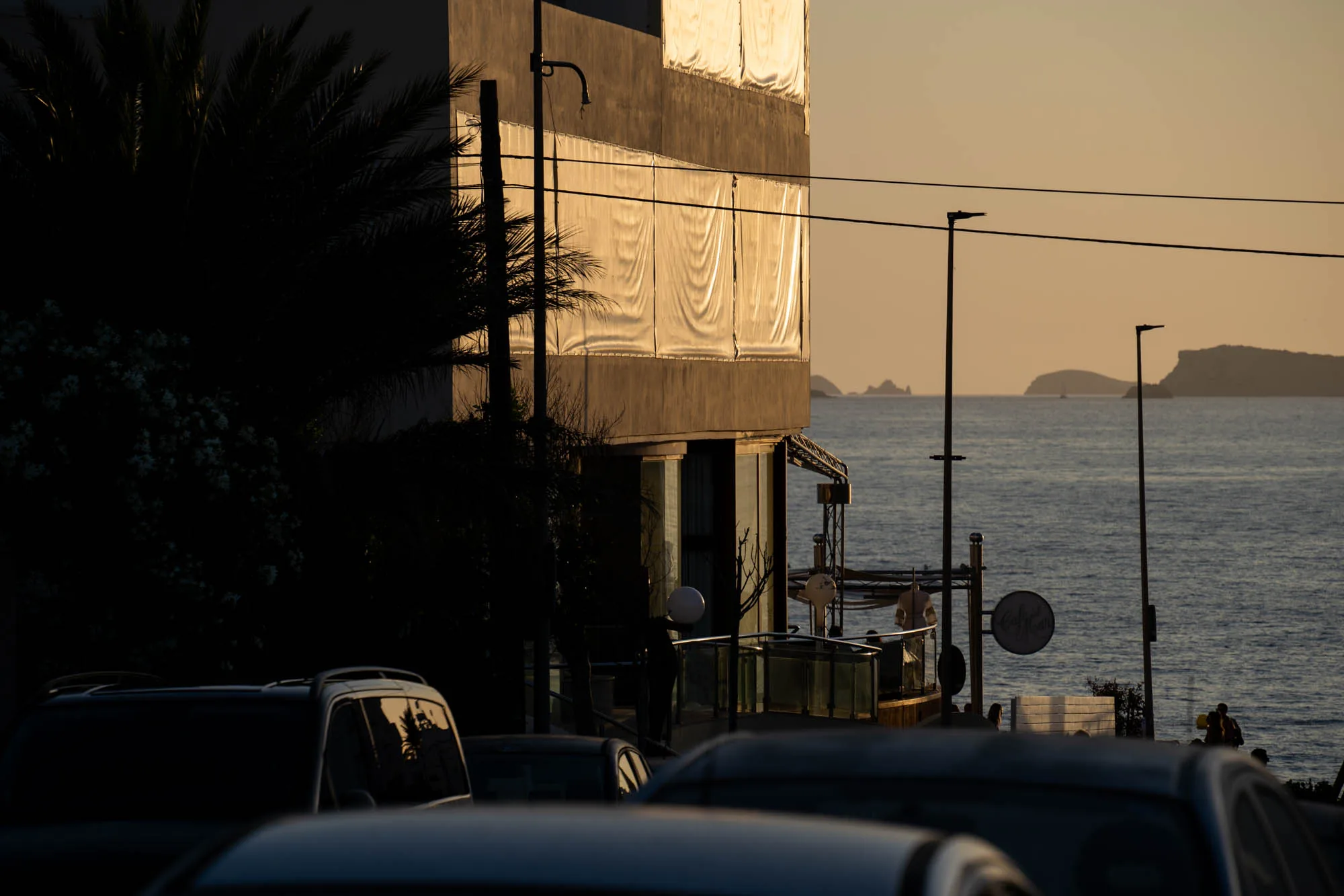 スペイン、イビサ島で撮影された夕暮れ時の写真です。前景には駐車場があり、複数の車の屋根が見えています。その奥には、ヤシの木や他の植物が生い茂り、一部は影になっていて詳細が見えません。建物の側面は白いシートのようなもので覆われており、夕日に照らされて金色に輝いています。背景には穏やかな海が広がり、水平線上にはいくつかの島影が見えます。電柱や電線が空を横切っており、人工的な要素も写真に加わっています。カフェのような場所も見え、人々が夕日を楽しんでいる様子が伺えます。全体的に落ち着いた雰囲気で、イビサ島の美しい夕景が捉えられています。