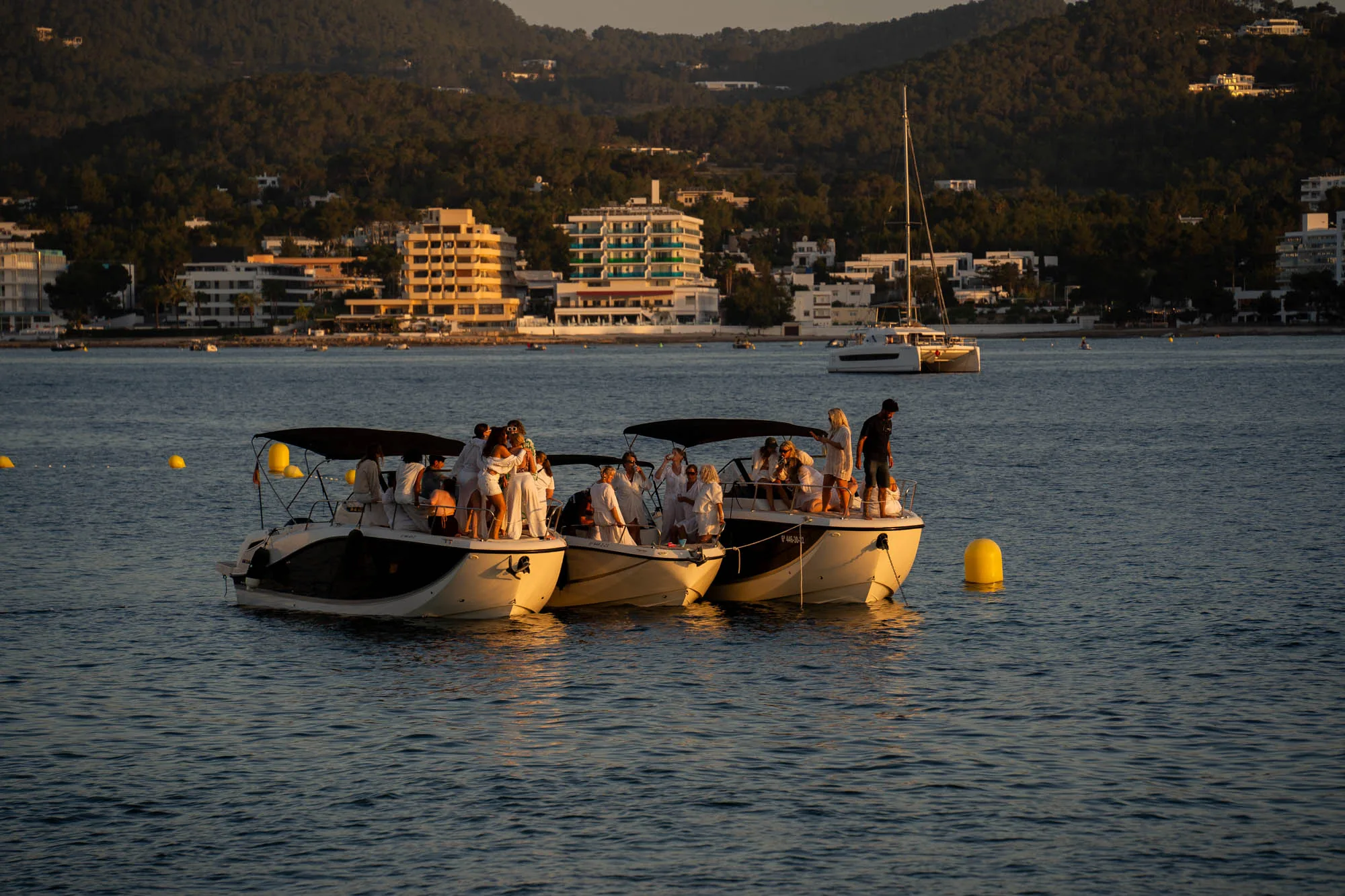 イビサ島の海で撮影された写真です。穏やかな海面に3隻のボートが並んで停泊しています。ボートの上には、白い服を着た人々が多数乗っており、パーティーの様子が伺えます。背景には、山と海岸沿いの建物群が見えます。夕暮れ時の光が全体を照らし、暖かく穏やかな雰囲気を醸し出しています。遠くには別の船が浮かんでいます。海にはボートを繋ぐ黄色のブイが浮かんでいます。