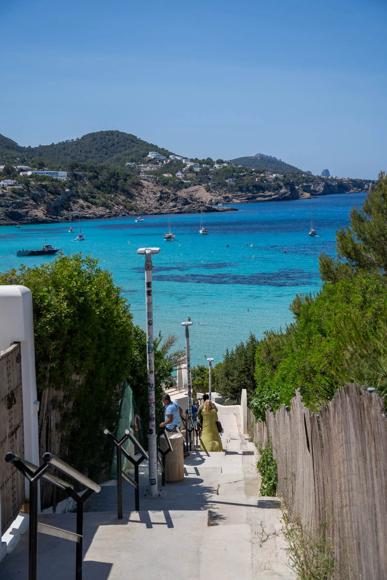 写真はスペインのイビサ島で撮影されたもので、階段を下る視点から海岸の風景が広がっています。ターコイズブルーの海は透明度が高く、遠浅になっているのがわかります。海にはヨットやボートが浮かび、海水浴を楽しむ人々も確認できます。海岸線は切り立った崖になっており、緑豊かな木々が生い茂っています。崖の上には白い家々が点在しており、リゾート地らしい雰囲気を醸し出しています。写真全体は明るく、夏の太陽が降り注いでいる様子が伝わってきます。階段の脇には手すりや街灯があり、生活感も感じられます。階段を下る人々の後ろ姿も写っており、これからビーチに向かうのか、あるいは帰る途中なのか、想像力を掻き立てられます。