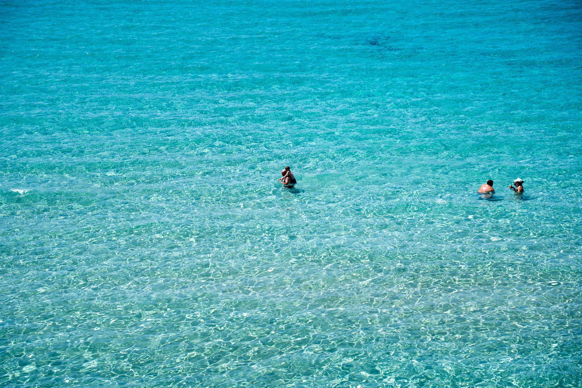 写真はスペインのイビサ島で撮影されたもので、透き通るようなターコイズブルーの海面が広範囲に写っています。水面は光を反射してきらきらと輝き、水の透明度が非常に高いことがわかります。海の中には、水着を着た数人の人物が写っています。一組は肩を組むようにして立っており、もう一組は少し離れた場所に並んで立っています。海水浴を楽しむ人々のリラックスした雰囲気が伝わってきます。全体的に、穏やかで美しいイビサの夏の日の情景を捉えた写真です。