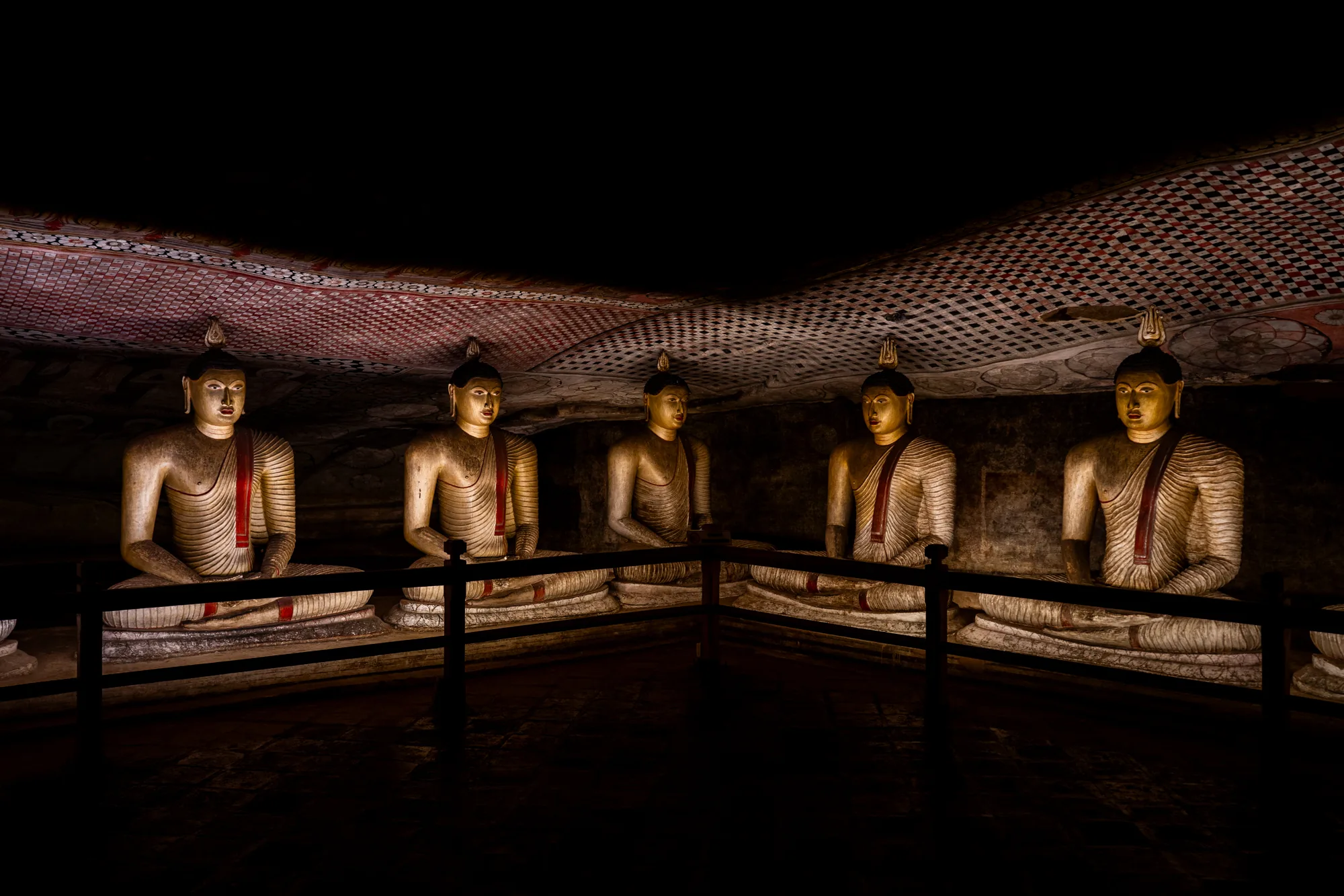 スリランカで撮影された写真には、5体の仏像が並んで座っている様子が写っています。仏像は、薄暗い洞窟のような空間に位置しており、それぞれが瞑想の姿勢をとっています。像は、白っぽい色合いで、赤いラインが袈裟のように表現されています。天井には、幾何学的な模様が描かれており、壁の一部にも同様の装飾が見られます。光は控えめで、仏像を照らし出すように当てられており、神秘的な雰囲気を醸し出しています。手前には、黒い柵のようなものが設置されており、仏像に近づけないようにしているようです。全体的に、宗教的な厳かさと静寂を感じさせる写真です。