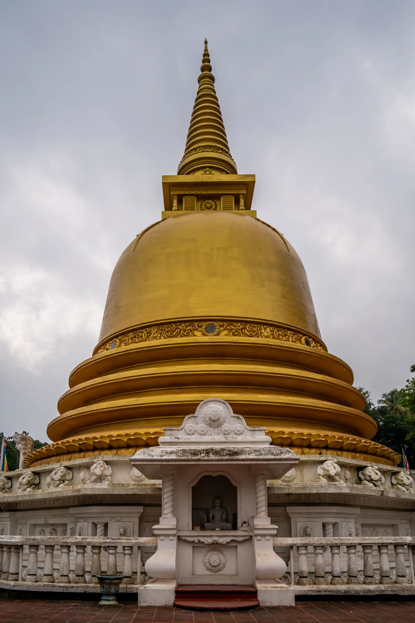 スリランカで撮影された写真には、黄金色に輝く仏塔が写っています。空は曇り空で、やや暗い印象です。仏塔はドーム状の本体の上に、円錐状の尖塔が重ねられた形状をしており、その表面は滑らかで光沢があります。本体部分には装飾的な模様が施され、細部まで丁寧に作られていることがわかります。仏塔の基壇部分は白く塗られており、小さな仏像が安置された祠が設けられています。基壇の周囲には、象の彫刻が並んでいます。地面は煉瓦色で舗装されており、建造物の荘厳さを引き立てています。全体的に落ち着いた雰囲気で、仏教寺院の神聖な雰囲気が伝わってくる写真です。