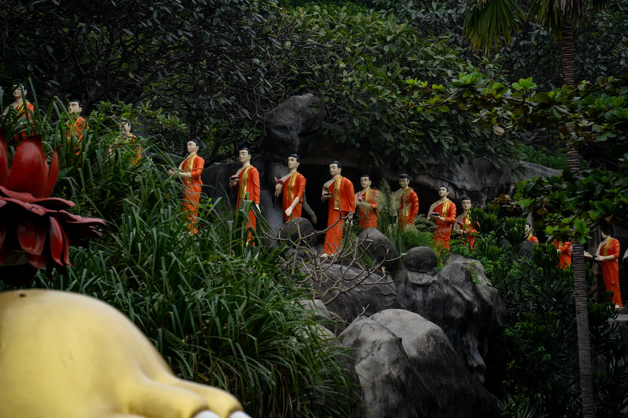 写真には、スリランカの緑豊かな風景の中に佇む仏像群が写っています。鮮やかなオレンジ色の衣をまとった仏像が、岩や植物が生い茂る斜面に沿って等間隔に並んでいます。それぞれの仏像は静かで穏やかな表情を浮かべ、手には蓮の花などを持っているようです。周囲は濃い緑の木々や草木に囲まれ、神秘的な雰囲気を醸し出しています。写真全体はやや暗めのトーンで、自然と仏教美術が調和した神聖な空間を表現しているようです。手前に見える大きな花や仏像の一部が、写真に奥行きを与えています。