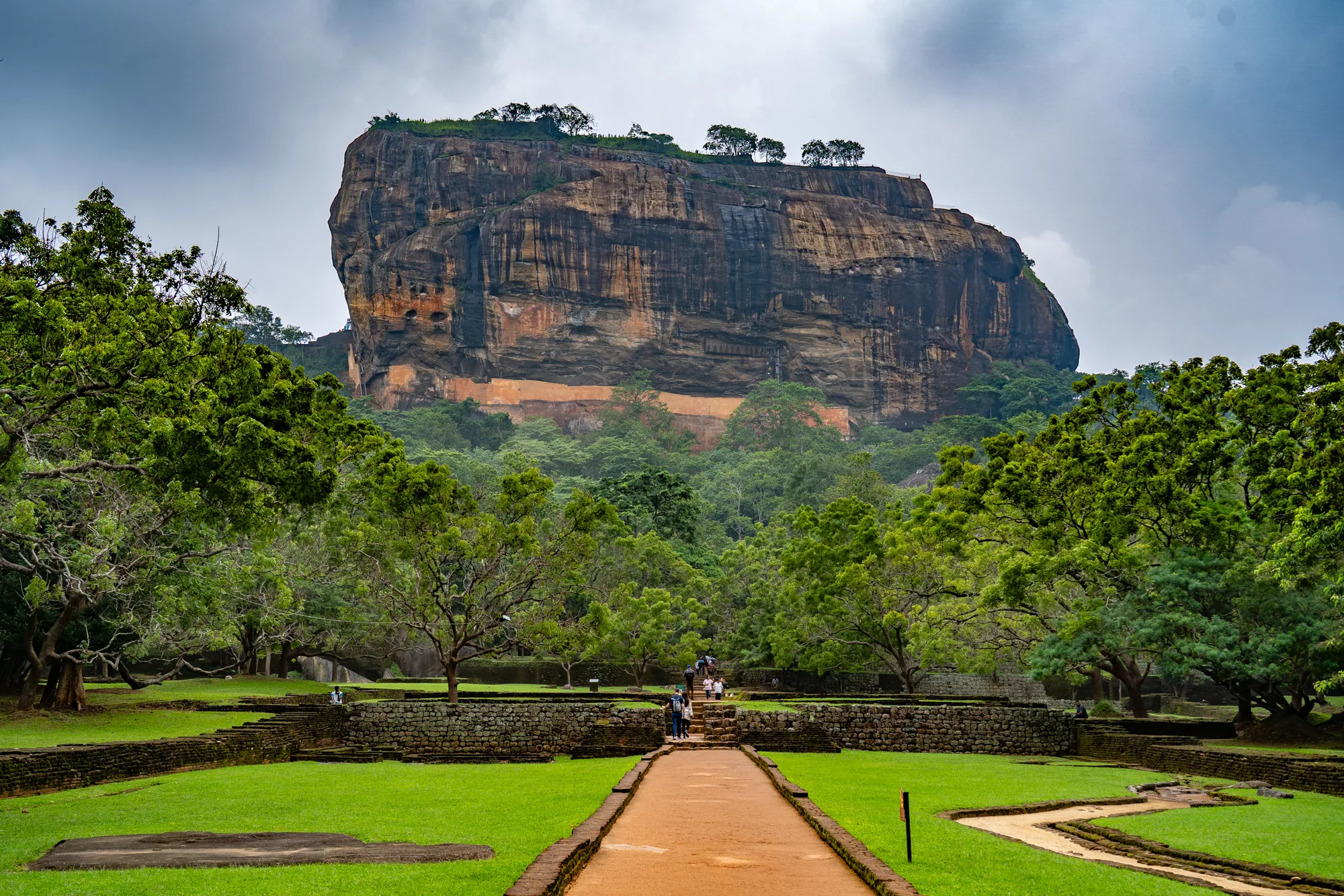 スリランカで撮影された風景写真です。画面奥には巨大な岩山であるシーギリヤロックがそびえ立っています。岩肌は垂直に切り立っており、茶色や黒色の縞模様が印象的です。頂上には木々が生い茂っているのが見えます。岩山の麓には緑豊かな木々が広がり、画面全体を覆っています。手前には整備された庭園があり、緑の芝生とレンガ造りの構造物が並んでいます。庭園の中央には、岩山へと続く道が伸びています。道には観光客と思われる人々の姿も見られます。空は曇っており、全体的に落ち着いた雰囲気です。歴史的な建造物と自然が調和した美しい景観が捉えられています。