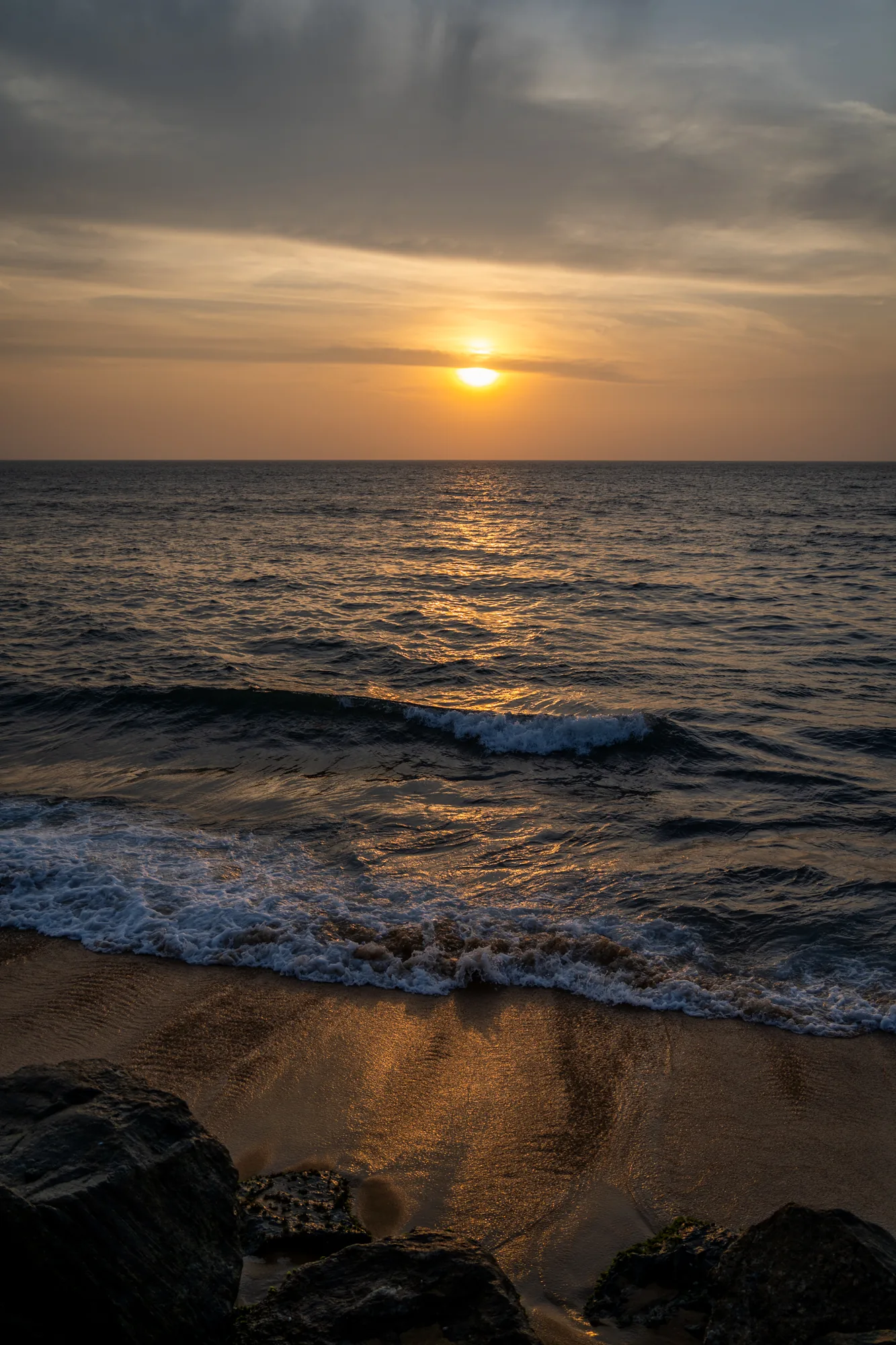 スリランカで撮影されたと思われる、美しい夕焼けの海の写真です。太陽は水平線に沈みかけており、空はオレンジ色とグレーの雲が混ざり合っています。太陽光は海面に反射し、キラキラと輝きを放っています。波は穏やかで、海岸に打ち寄せています。手前には黒い岩がいくつか配置されており、写真に奥行きを与えています。全体的に、穏やかでリラックスできる雰囲気の写真です。太陽の光が反射している様子が印象的で、美しい情景を切り取っています。