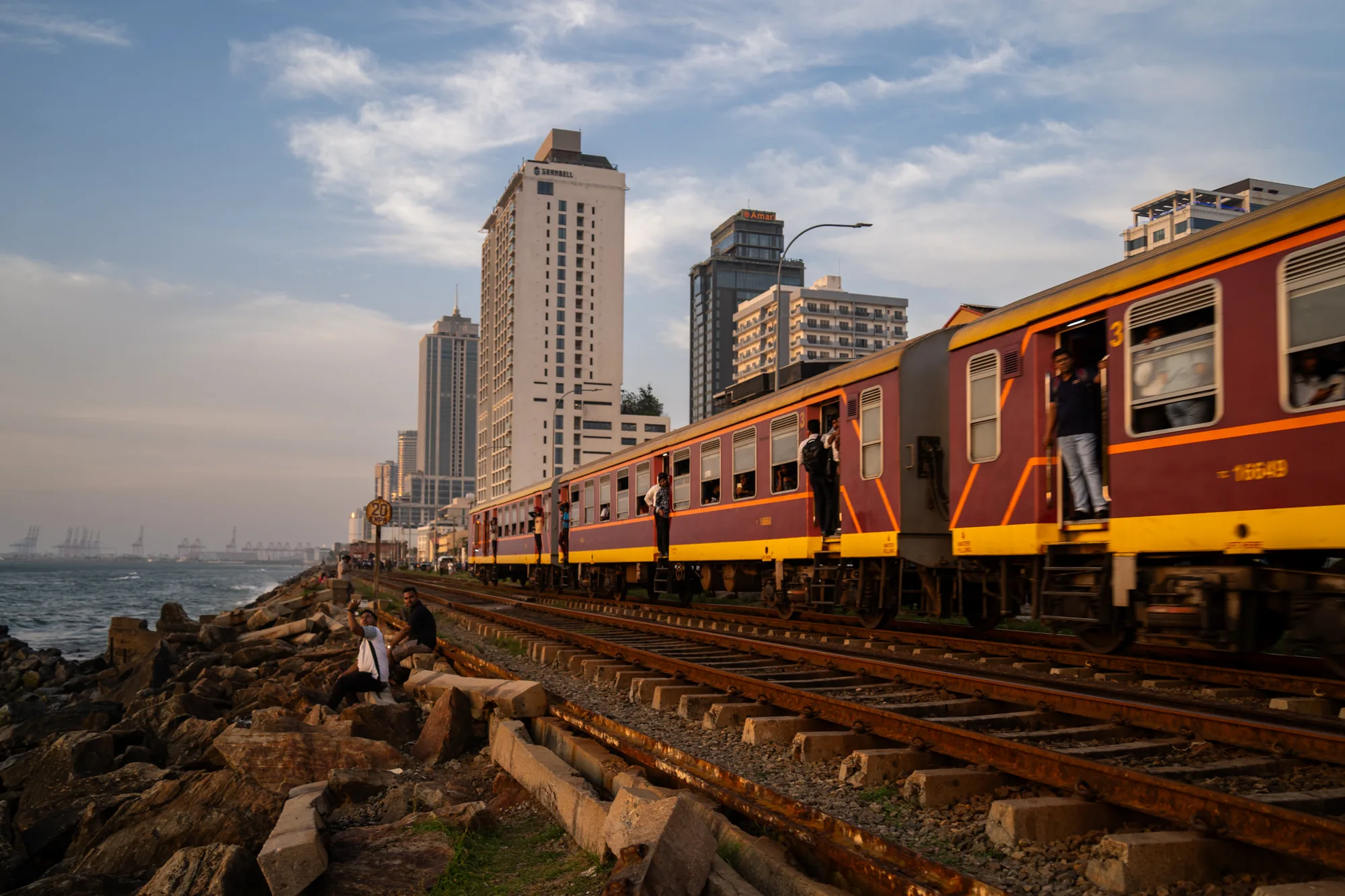 写真は、スリランカの海沿いの風景を捉えたものです。画面右側には、エンジ色と黄色を基調とした列車が走行しており、多くの乗客が窓から顔を出したり、ドア付近に立っていたりしています。列車の窓枠には装飾的なデザインが見られます。列車は右から左へと動いており、背景には複数の高層ビルがそびえ立っています。空は青空で、薄い雲が浮かんでいます。  画面左側には、海岸線があり、岩が積み上げられています。その岩の上に、数人の人々が座って景色を眺めたり、写真を撮ったりしています。遠くには、港湾施設のようなものが見え、クレーンが並んでいる様子も確認できます。全体的に、夕暮れ時の柔らかい光が差し込み、穏やかでノスタルジックな雰囲気を醸し出しています。列車の活気と、海岸の静けさが対照的な、スリランカの日常風景を切り取った一枚と言えるでしょう。