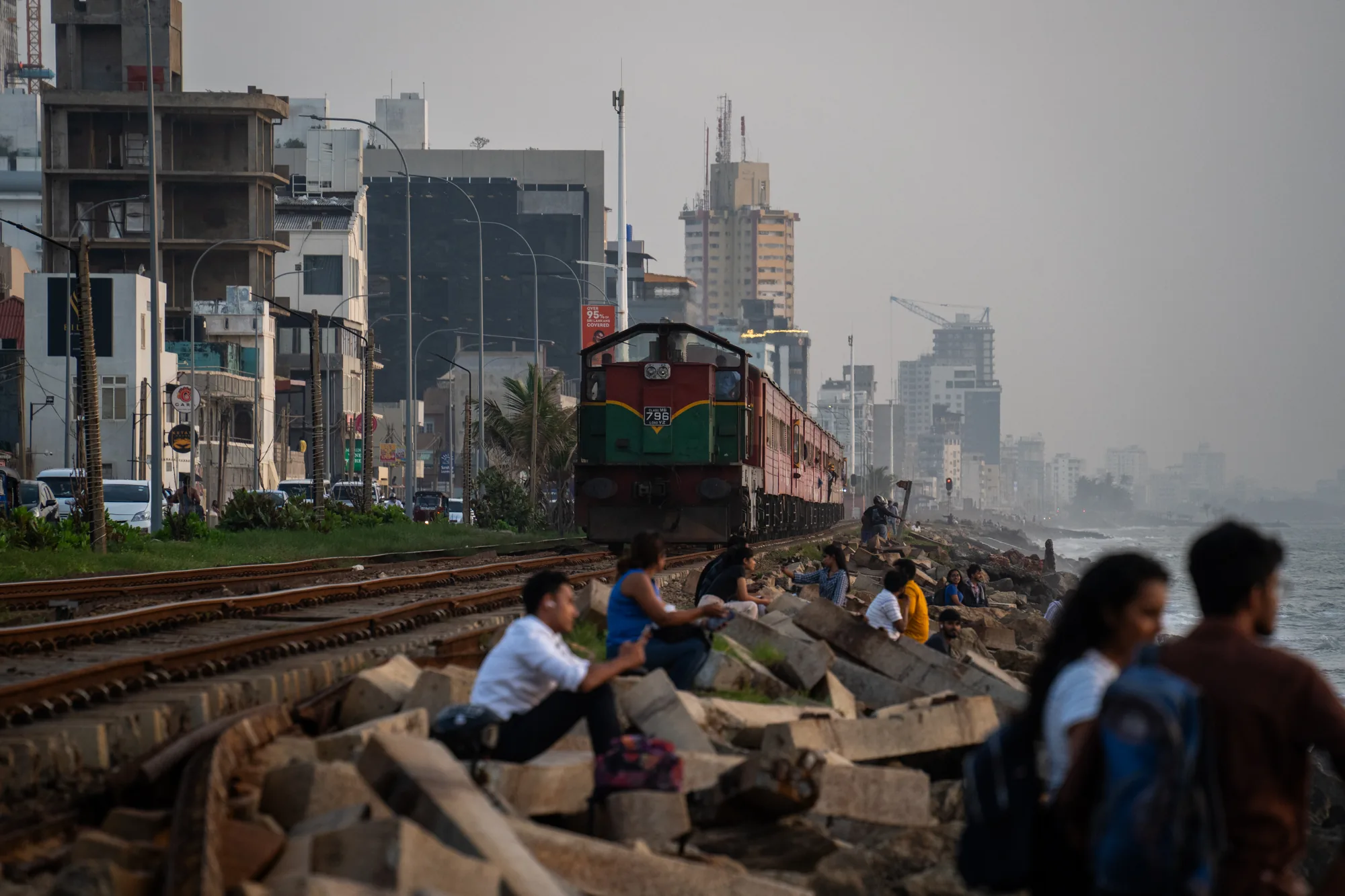 スリランカで撮影された写真には、海岸線に沿って走る列車が写っています。手前にはコンクリートの塊が積み上げられており、そこに人々が腰掛けて海を眺めています。列車のすぐ脇にも人が座っており、夕暮れ時の穏やかな時間を楽しんでいるようです。遠景には高層ビル群が立ち並び、開発が進んでいる様子が伺えます。空はやや霞んでおり、全体的に落ち着いた雰囲気の写真です。線路沿いには緑も生い茂っており、都市と自然が共存している様子が感じられます。