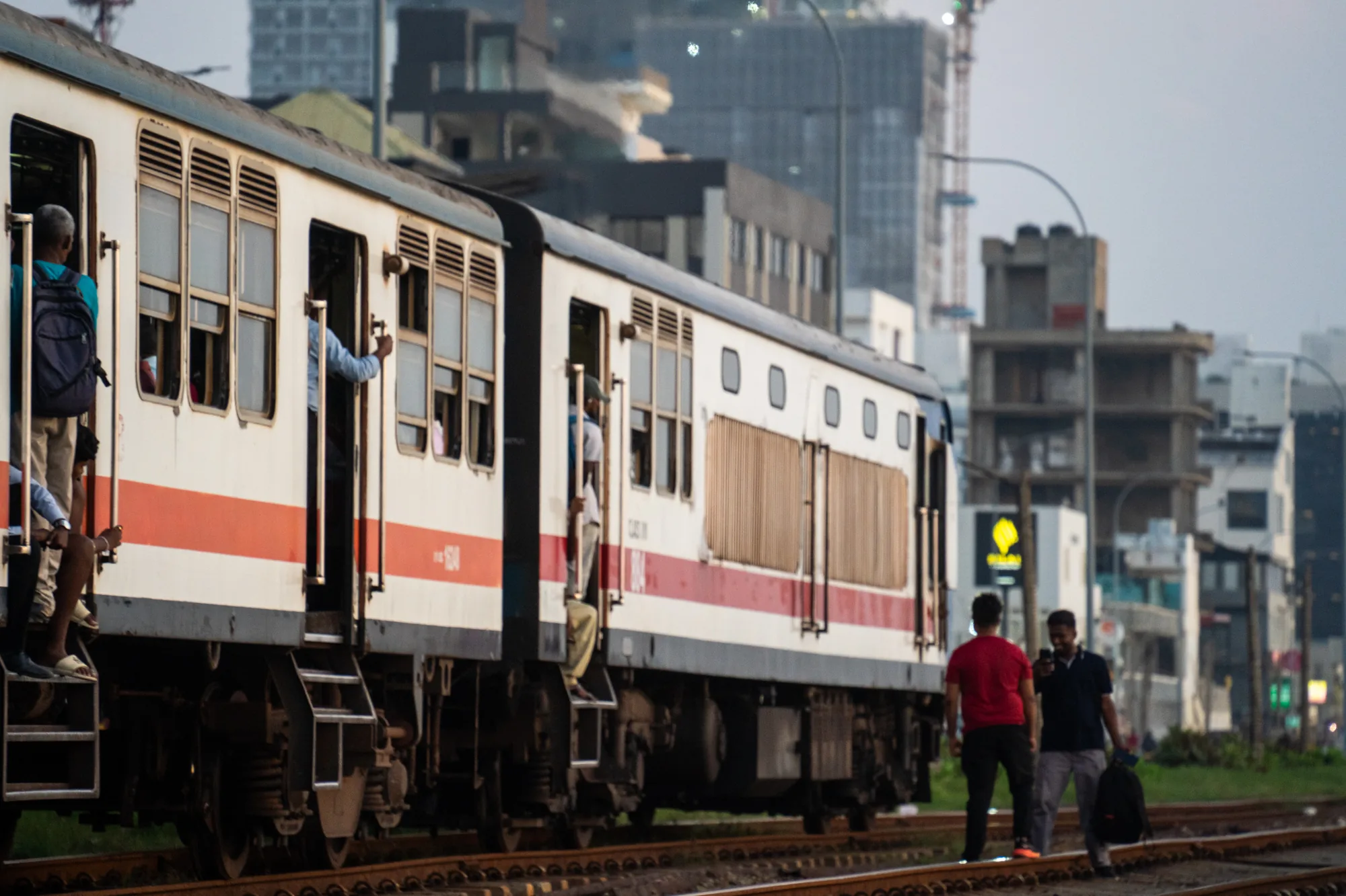 スリランカで撮影された写真です。手前には線路が走り、そこに白い車体に赤いラインが入った列車が写っています。列車のドアは開け放たれており、何人かの人がドア付近に立っているのが見えます。乗客の中には、ドアから足を投げ出して座っている人もいます。列車は走行中なのか停車中なのかは不明です。背景には都市部の建物が立ち並び、建設中のクレーンも見えます。線路の脇には草が生い茂り、若い男性2人が線路の上を歩いています。一人は赤いシャツを着ており、もう一人は黒いシャツを着てリュックを背負っています。全体的に、スリランカの日常的な通勤風景を切り取った写真と言えるでしょう。やや霞がかった空や、夕暮れ時のような光の加減から、どこかノスタルジックな雰囲気も感じられます。