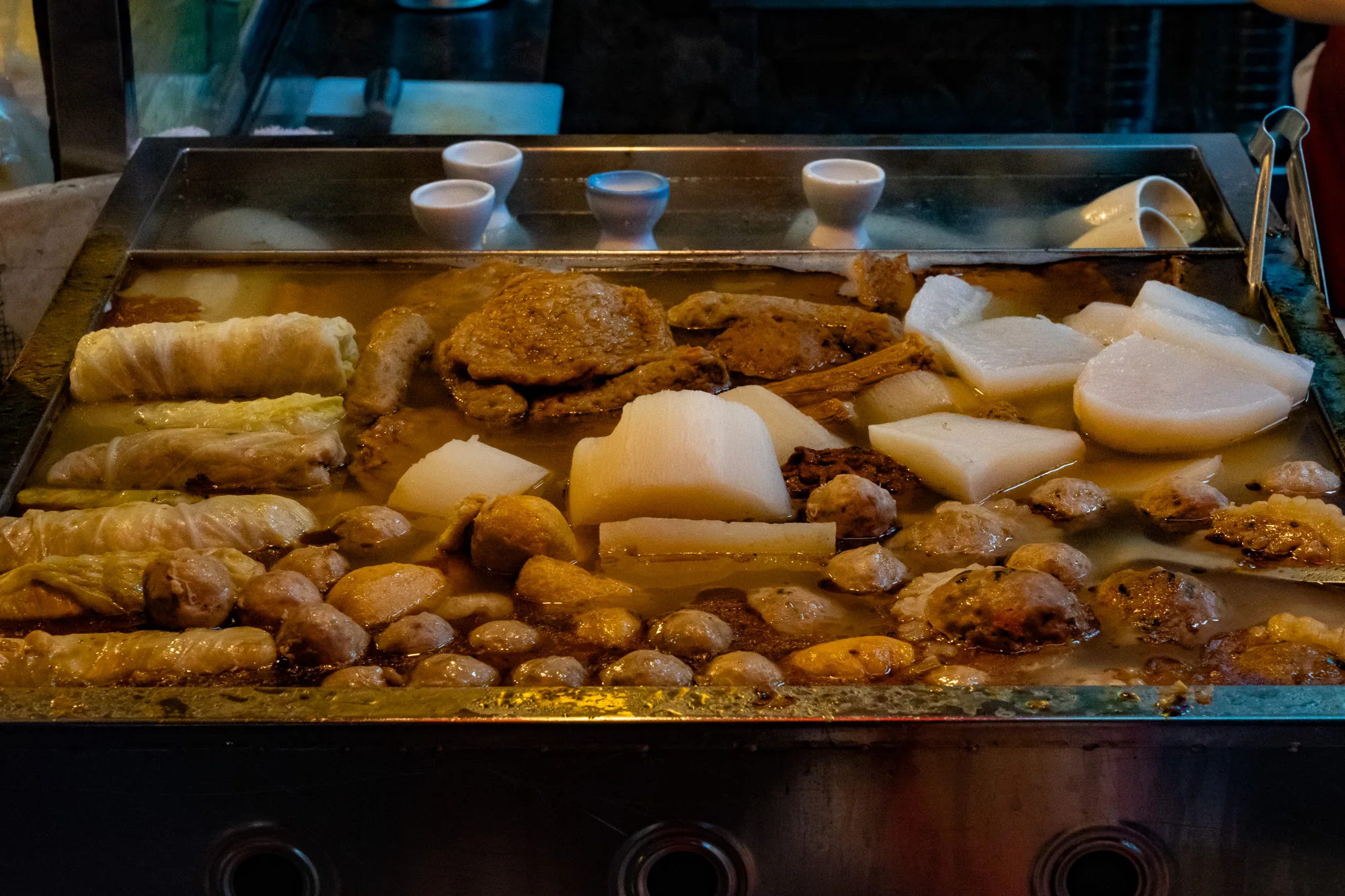 台湾の台南で撮影された写真です。屋台のおでん鍋が写っており、様々な具材が煮込まれています。白菜巻き、ミートボール、大根、練り物などが見えます。スープは茶色で、具材に味が染み込んでいる様子が伺えます。鍋の上には白い小さな器が並べられています。屋台の照明が具材を照らし、温かみのある雰囲気です。夜市の食べ歩きを連想させる、食欲をそそる一枚です。