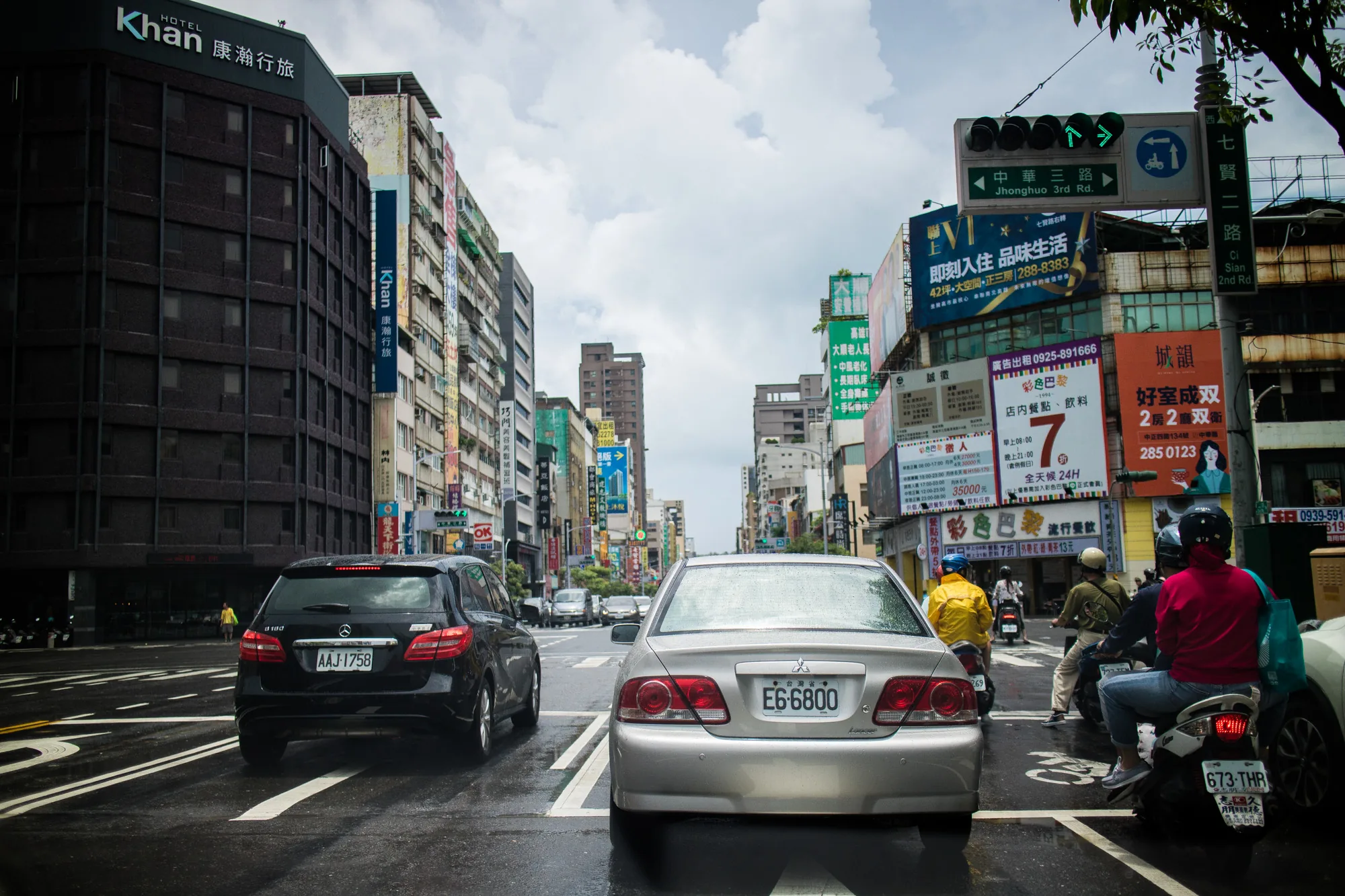 写真は台湾の台南で撮影された、雨上がりの街の風景です。画面中央には銀色の乗用車が停車しており、リアライトが濡れた路面に反射しています。車のナンバープレートには「E6-6800」と書かれており、「台灣省」の文字も見えます。車の背後には、複数台のバイクが停車しており、運転手や同乗者が信号待ちをしている様子が伺えます。  道路の向こうには、高層ビルが立ち並ぶ街並みが広がっています。「Khan HOTEL 康瀚行旅」という看板を掲げたホテルや、広告看板が数多く見られます。看板には中国語の文字が書かれており、様々な情報が伝えられています。空には雲が広がり、天気は曇りのようです。路面は濡れており、雨上がりの湿った空気感が伝わってきます。全体的に、台湾の活気ある都市の日常を切り取ったような写真です。