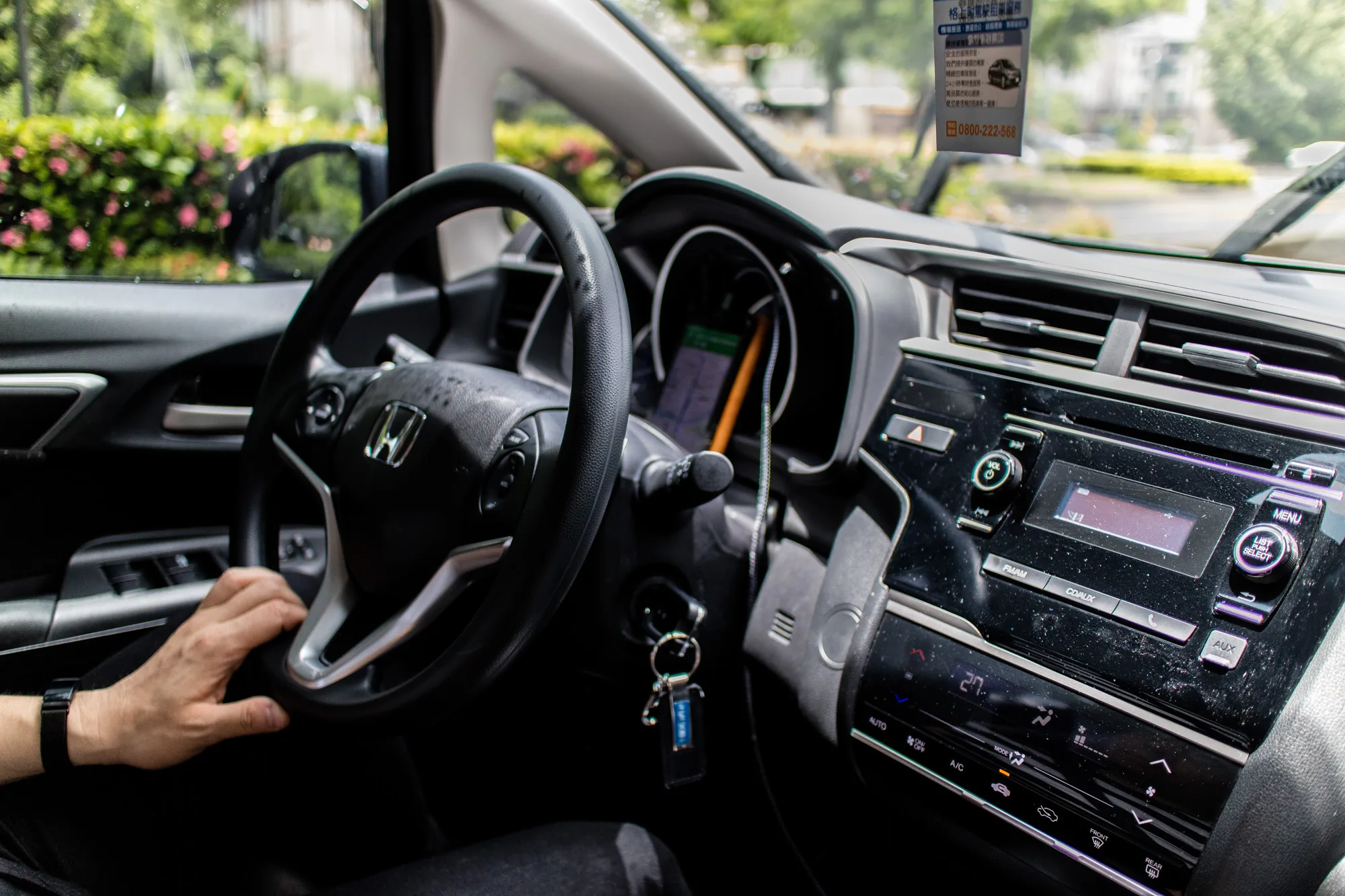 この写真は台湾の台南で撮影されたもので、車の運転席からの視点を捉えています。運転席の様子が中心に写っており、運転手の左手がステアリングを握っているのが見えます。手首には黒い腕時計が装着されています。ステアリングの奥にはホンダのエンブレムがあり、車種を特定することができます。  ダッシュボードは黒色で、カーナビと思われる機器やオーディオシステム、エアコンの操作パネルなどが配置されています。カーナビには地図が表示されているようです。フロントガラスには雨が当たっており、ワイパーが作動していることが分かります。また、格上附駕駛租車服務（運転手付きレンタカーサービス）の広告がフロントガラスに貼り付けられています。  車の外には緑豊かな風景が広がっており、ピンク色の花が咲いている低木も見えます。全体的に、雨の中のドライブシーンを切り取った写真であり、車内からの落ち着いた雰囲気が伝わってきます。