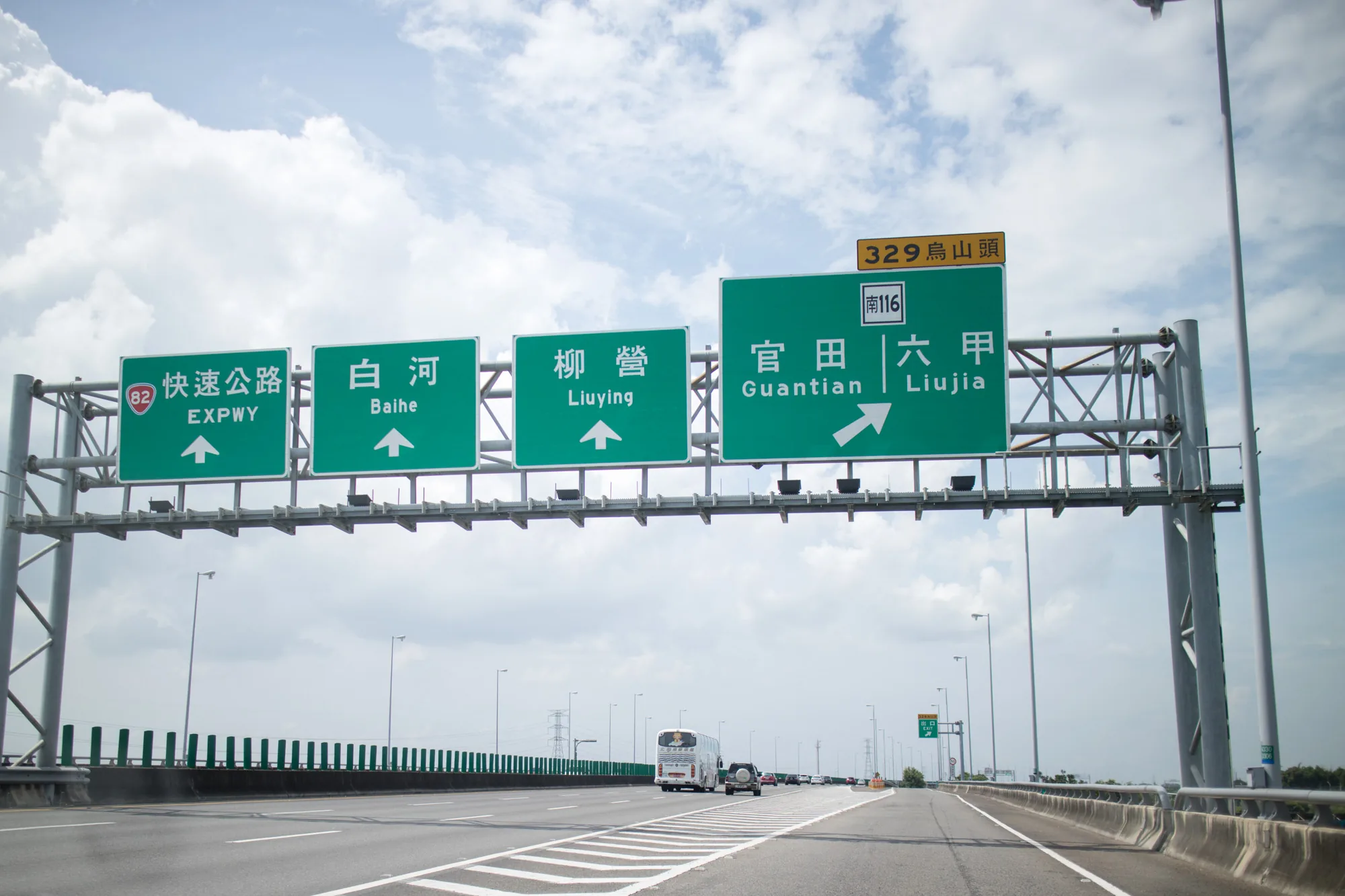 写真は台湾の台南で撮影された高速道路の風景です。画面上部には、緑色の大きな案内標識が設置されています。標識には、高速道路82号線(EXPWY)、白河(Baihe)、柳營(Liuying)、官田(Guantian)、六甲(Liujia)といった地名が、それぞれ矢印と共に表示されています。その上には「329 烏山頭」と小さく書かれた標識もあります。高速道路上には数台の車両が走行しており、白いバスやSUVのような車が見えます。道路はアスファルトで舗装されており、白線が引かれています。背景には曇り空が広がっており、電柱や緑色の防護柵も見えます。全体的に、高速道路を走行中に撮影された写真であり、道路標識が明確に写っています。