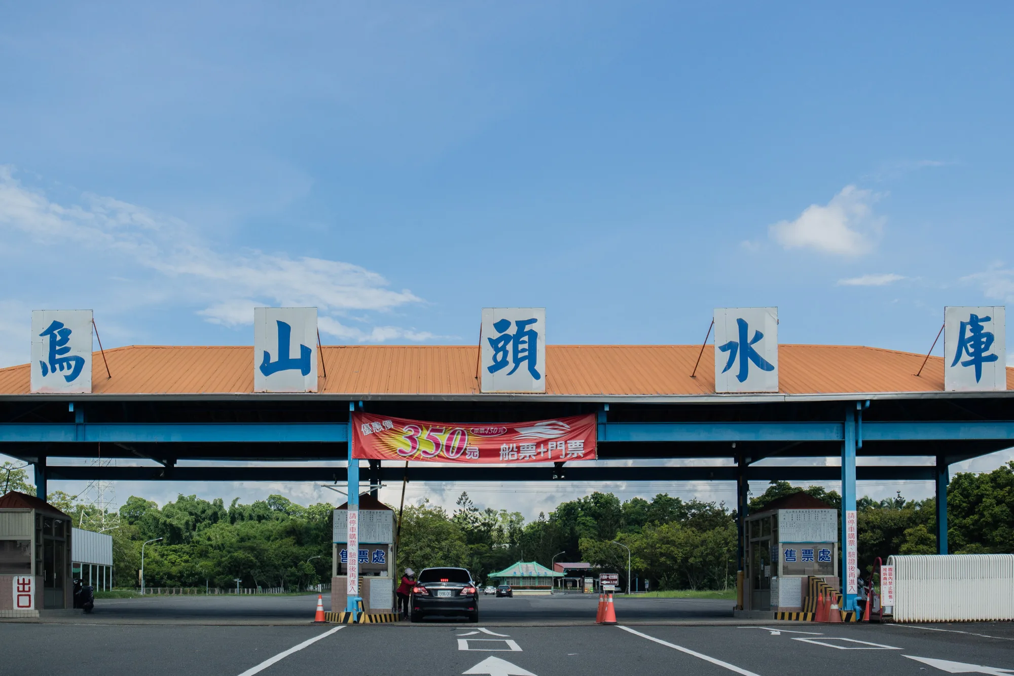 写真は台湾の台南にある烏山頭水庫の入口を捉えています。青い空が広がり、ゲートの屋根には「烏山頭水庫」と書かれた白い看板が青い文字で掲げられています。ゲート自体は青い鉄骨で組まれ、屋根はオレンジ色をしています。ゲートの上部には、「優惠價 350 元 原價450元 船票+門票」(特別価格350元 通常価格450元 船チケット+入場料)と書かれた赤い横断幕が掲げられています。ゲートの手前には、黒い車が停車しており、料金所でチケットを購入している様子が見えます。ゲートの左右には料金所ブースがあり、白い柵が設置されています。アスファルトの道路には白い矢印が描かれており、進む方向を示しています。背景には緑豊かな木々が茂り、公園のような雰囲気を感じさせます。全体的に、観光地である烏山頭水庫の明るく開放的なイメージが伝わってくる写真です。