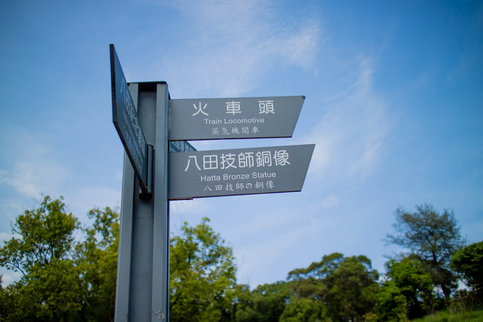 台湾の台南で撮影されたと思われる写真には、観光地の案内標識が写っています。標識は灰色の金属製で、いくつかの方向を示す矢印の形をしたプレートが取り付けられています。一番上のプレートには「火車頭」と書かれ、その下に英語で「Train Locomotive」、さらにその下に「蒸気機関車」と書かれています。次のプレートには「八田技師銅像」と書かれ、その下に英語で「Hatta Bronze Statue」、さらにその下に「八田技師の銅像」と書かれています。標識の背景には、青空と白い雲が広がっており、緑豊かな木々が茂っています。日中の屋外で撮影された写真で、晴れた日の風景を捉えています。