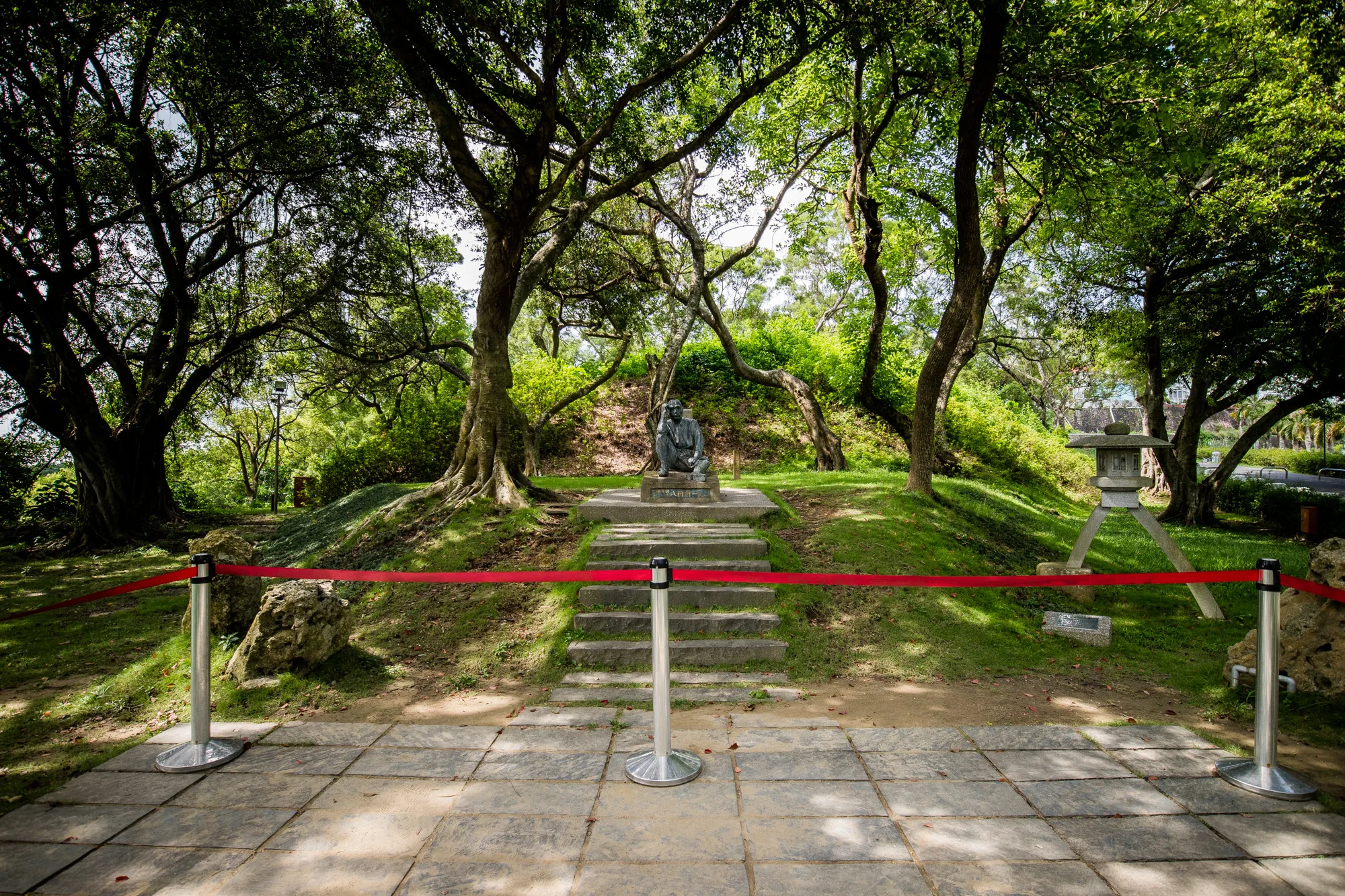 写真は台湾の台南で撮影された、緑豊かな公園の一角を捉えています。前景は正方形の石畳の道で、道の奥には銀色のポールと赤いロープが張られ、その先への立ち入りを制限しているようです。  ロープの奥は緑の芝生が広がり、緩やかな傾斜の上に、石段があります。石段を登った先には、思索にふける人物の像が鎮座しています。像の周囲は、根を張った大きな木々が茂り、木漏れ日が像を照らしています。  写真右側には、石灯籠が見えます。背景にはさらに緑が生い茂り、遠くまで緑豊かな風景が広がっていることがうかがえます。全体的に静かで落ち着いた雰囲気の写真です。