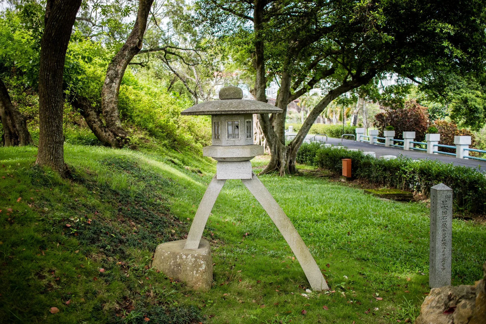 台湾の台南で撮影された写真には、緑豊かな公園の風景が広がっています。中央には、石造りの灯籠が置かれています。灯籠は笠、火袋、中台、基礎から構成されており、歴史を感じさせる風合いです。灯籠の足は斜めに伸び、地面に固定されています。背景には緑の芝生と木々が生い茂り、自然豊かな様子が伺えます。奥には遊歩道のようなものが見え、市民の憩いの場となっているようです。灯籠の隣には石碑が立っており、日本語の文字が刻まれていることから、日本との関わりがある場所であることが推測されます。全体的に穏やかで静かな雰囲気が感じられる写真です。