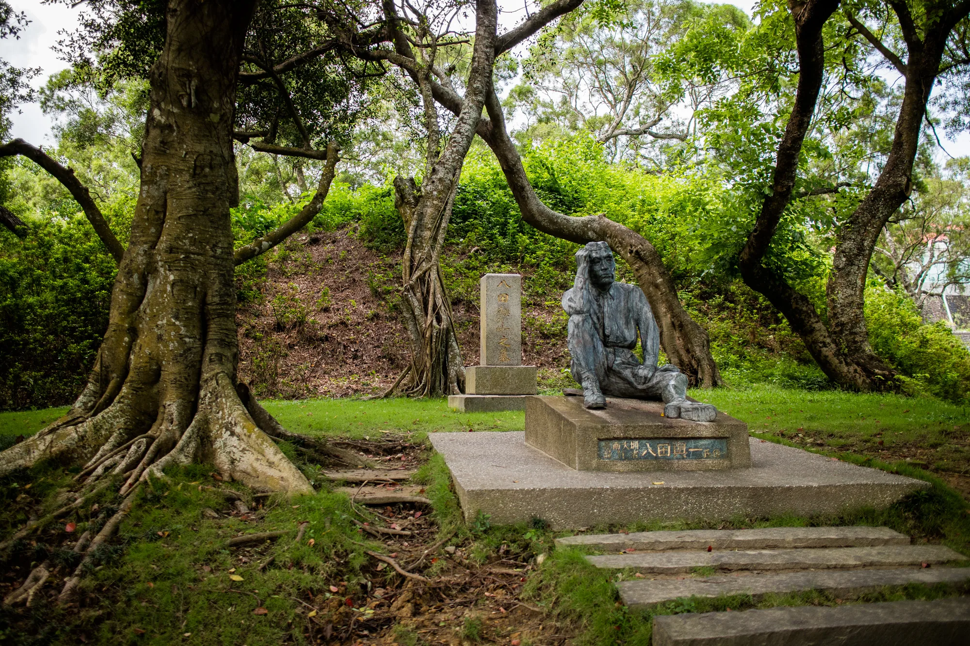 台湾の台南で撮影された写真です。鬱蒼とした緑に囲まれた場所に、石造りの台座の上に座り込む人物の像が置かれています。像は男性で、頭に手を当てて物思いにふけっているようなポーズをとっています。像の隣には、文字が刻まれた石碑が立っています。周囲は木々が生い茂り、苔むした石段が像へと続いています。全体的に静かで落ち着いた雰囲気で、歴史を感じさせる場所であることが伺えます。木漏れ日が差し込み、神秘的な雰囲気を醸し出しています。