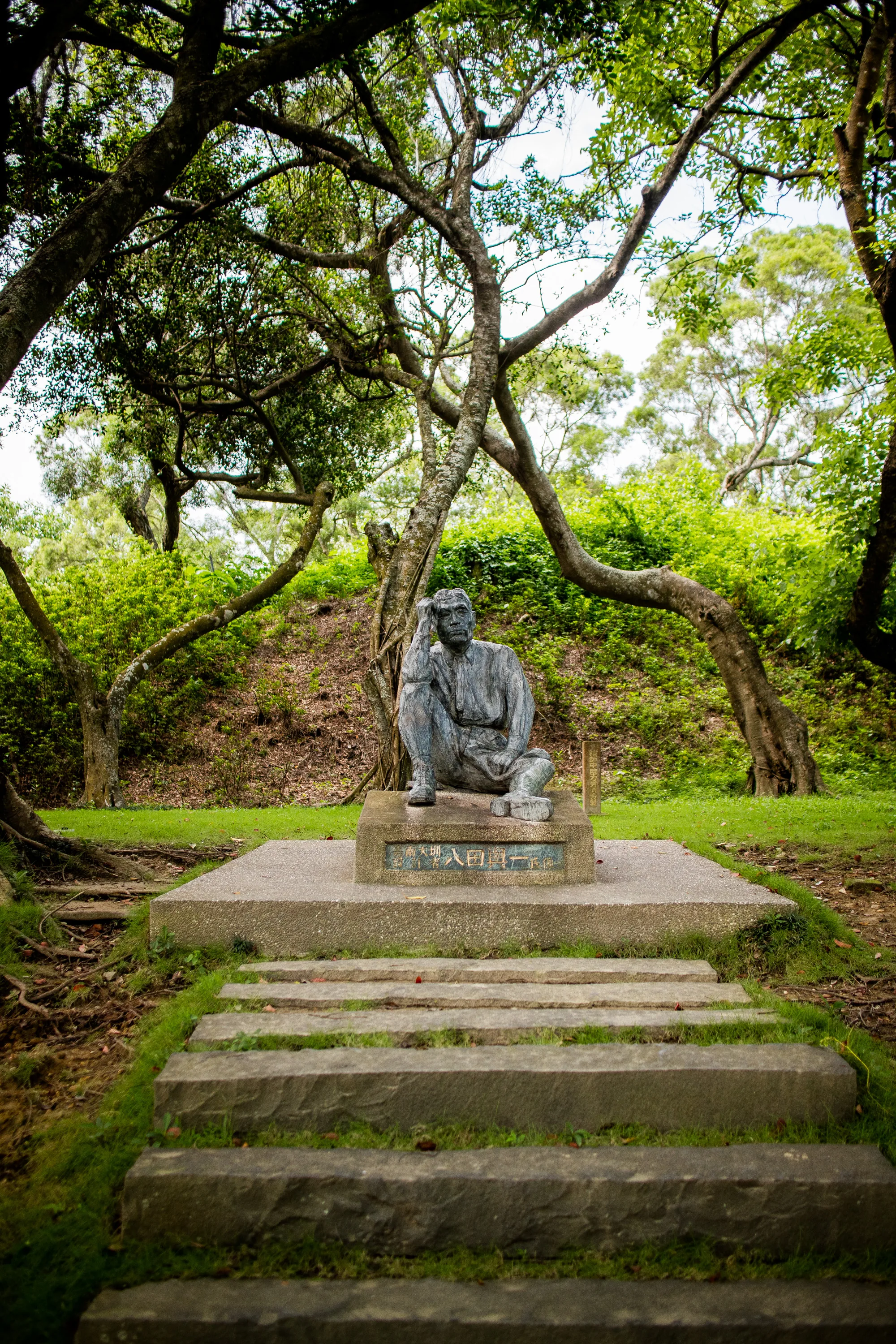 この写真は台湾の台南で撮影されたもので、緑豊かな木々に囲まれた場所にある人物像を捉えています。画面手前には、苔むした石段が数段設けられており、その先にはコンクリート製の台座が見えます。台座の上には、座った姿勢で物思いにふけるような表情を浮かべた人物の像が置かれています。像は服を着ており、頭に手を当てています。台座には漢字が刻まれています。背景には緑が生い茂り、木々の枝が空を覆うように広がっています。全体的に静かで落ち着いた雰囲気が漂っており、歴史的な場所や公園の一角を切り取ったような印象を受けます。