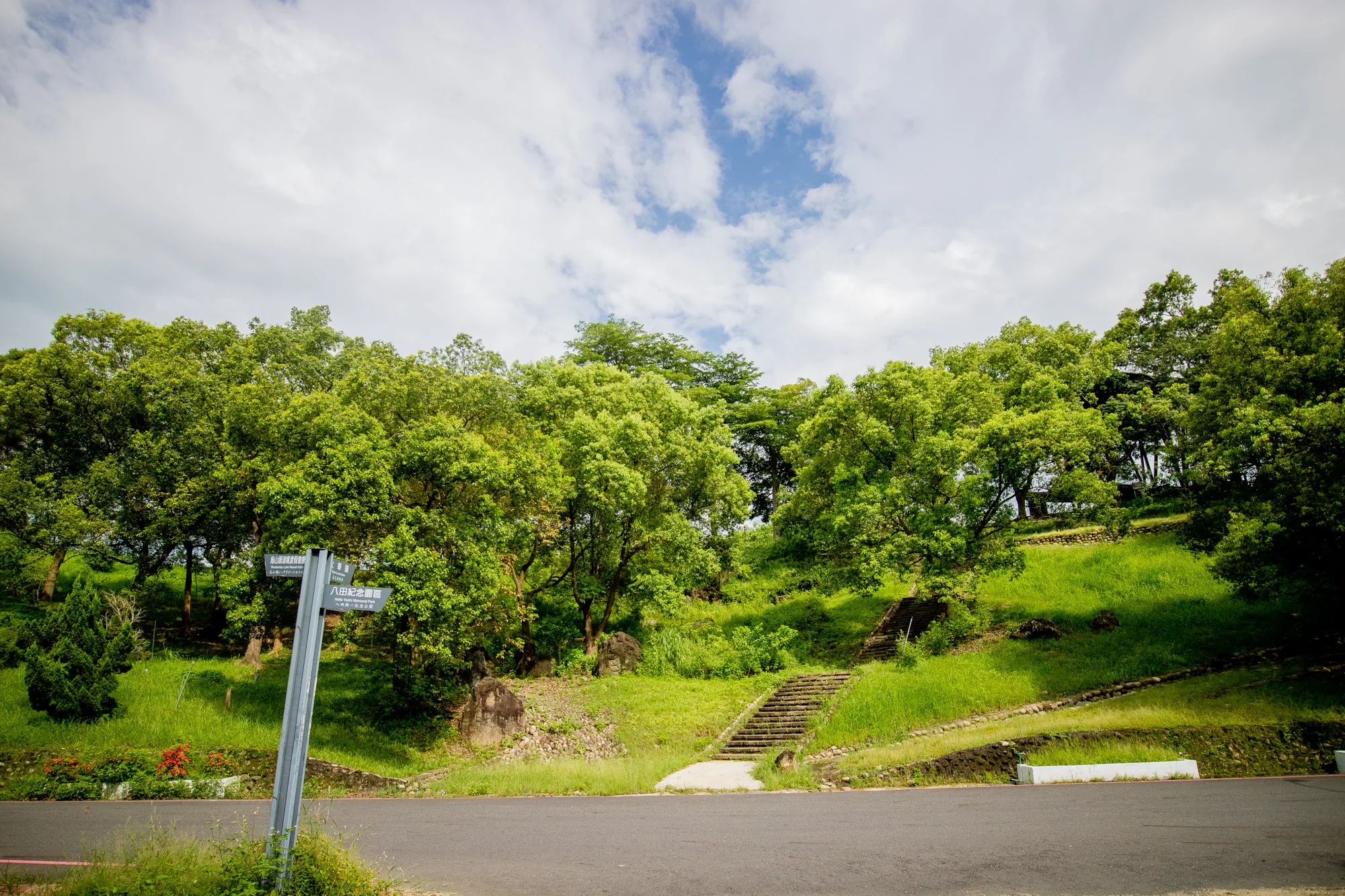 写真は台湾の台南で撮影された、緑豊かな公園の風景です。画面の下部はアスファルトの道路で占められており、その先には緑色の斜面が広がっています。斜面には木々が生い茂り、空には白い雲が浮かんでいます。斜面には階段があり、上へと続いています。左側には、観光地への方向を示す案内標識が立っています。全体的に、穏やかで自然豊かな雰囲気の画像です。