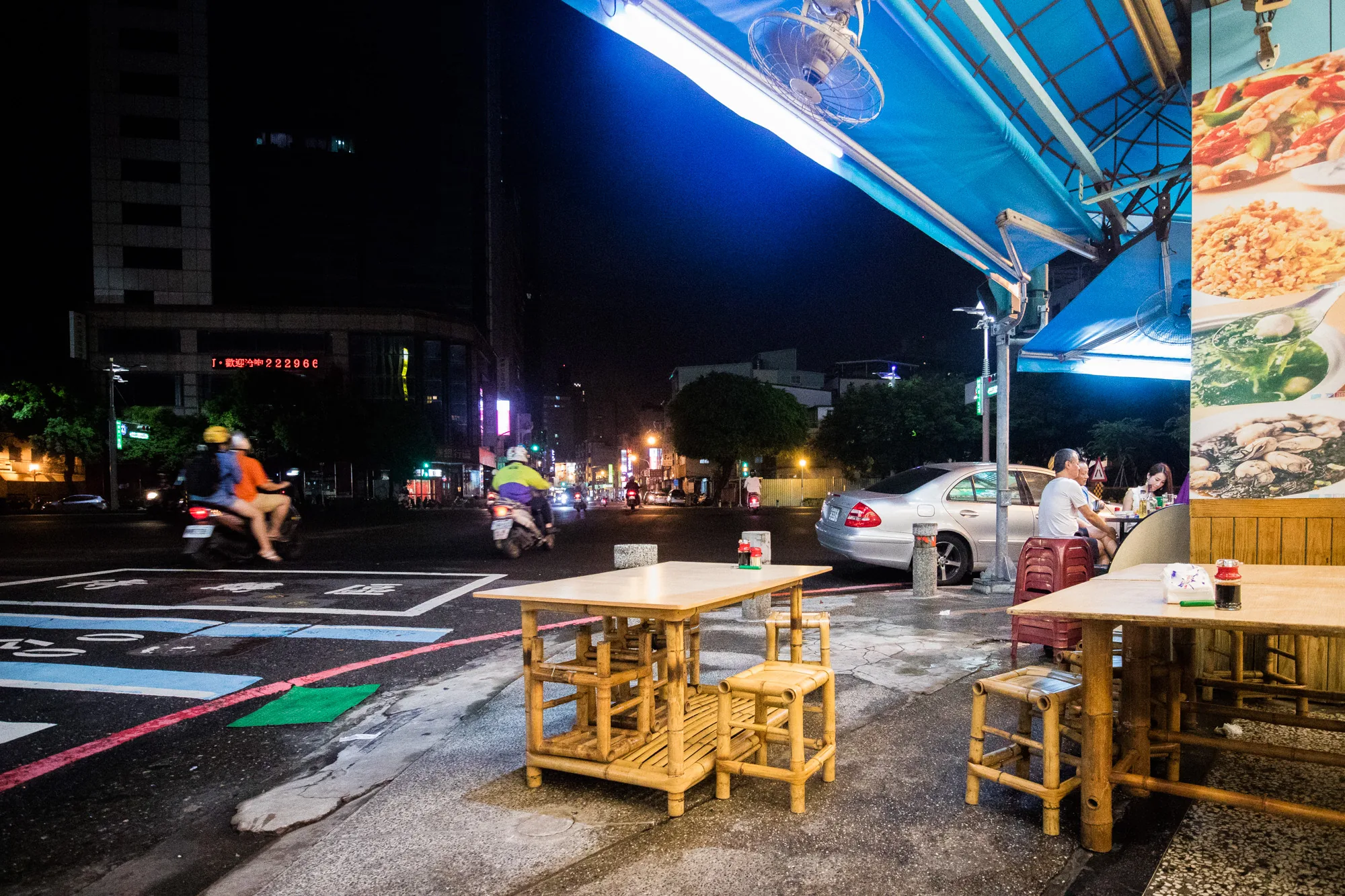 台湾の台南で撮影された夜の写真です。路上に置かれた竹製のテーブルと椅子が手前に配置されており、台湾らしい屋台の雰囲気を醸し出しています。テーブルの上には調味料などが置かれ、食事の準備がされているようです。背景には交通量の多い道路があり、バイクが走っています。歩道には食事をしている人々が写っており、地元の人が夜の屋台を楽しんでいる様子が伝わってきます。お店の看板には料理の写真が大きくプリントされており、食欲をそそります。夜空は暗く、街灯の光が温かみを加えています。全体的に活気があり、親しみやすい雰囲気の写真です。