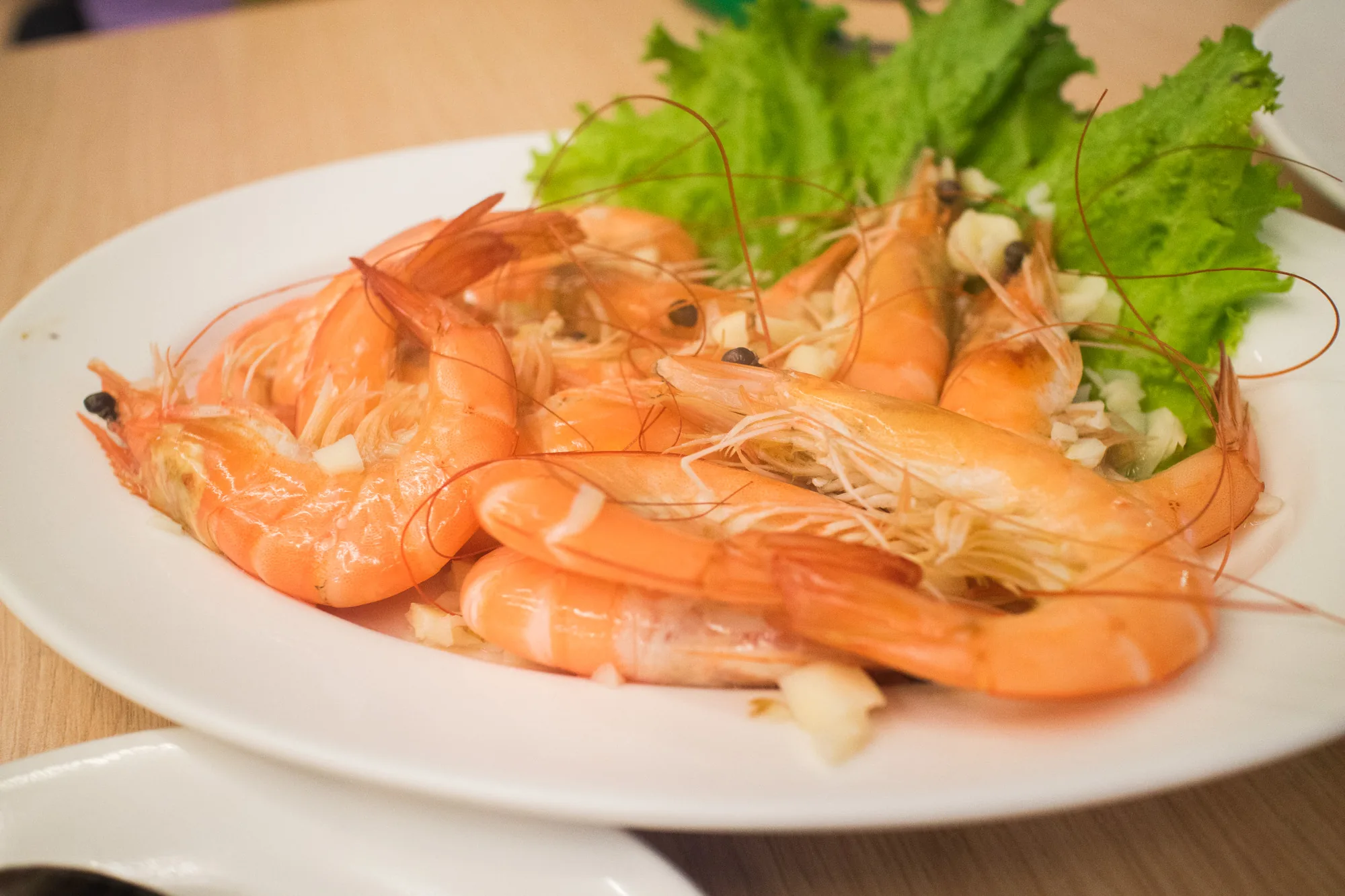 写真には、白くて丸い皿に盛られたエビのニンニク蒸しが写っています。エビは鮮やかなオレンジ色をしており、殻付きのまま調理されているようです。エビの上には細かく刻まれたニンニクが散りばめられています。皿の奥には、鮮やかな緑色のレタスが添えられています。背景はぼやけていますが、木製のテーブルの上に皿が置かれていることが分かります。照明は明るく、料理の鮮やかさを引き立てています。全体的に食欲をそそる、美味しそうな写真です。