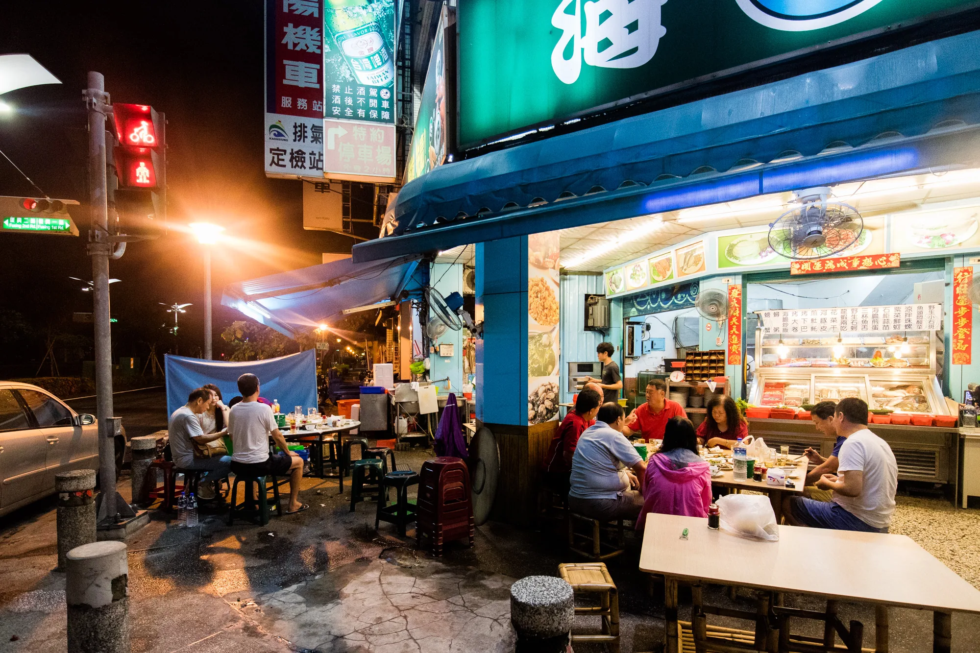 写真は、台湾の台南で撮影された夜の屋台の様子を捉えています。通りに面した屋台には、テーブル席がいくつか設けられており、人々が食事を楽しんでいます。屋台の看板には中国語が書かれており、様々な料理の写真が飾られています。店内には料理が並べられたショーケースがあり、店員が調理をしている様子も伺えます。  道路は濡れており、夜の街灯が反射して光っています。奥にはバイクの絵柄のある信号機が見えます。 全体的に賑やかで活気のある雰囲気の写真です。