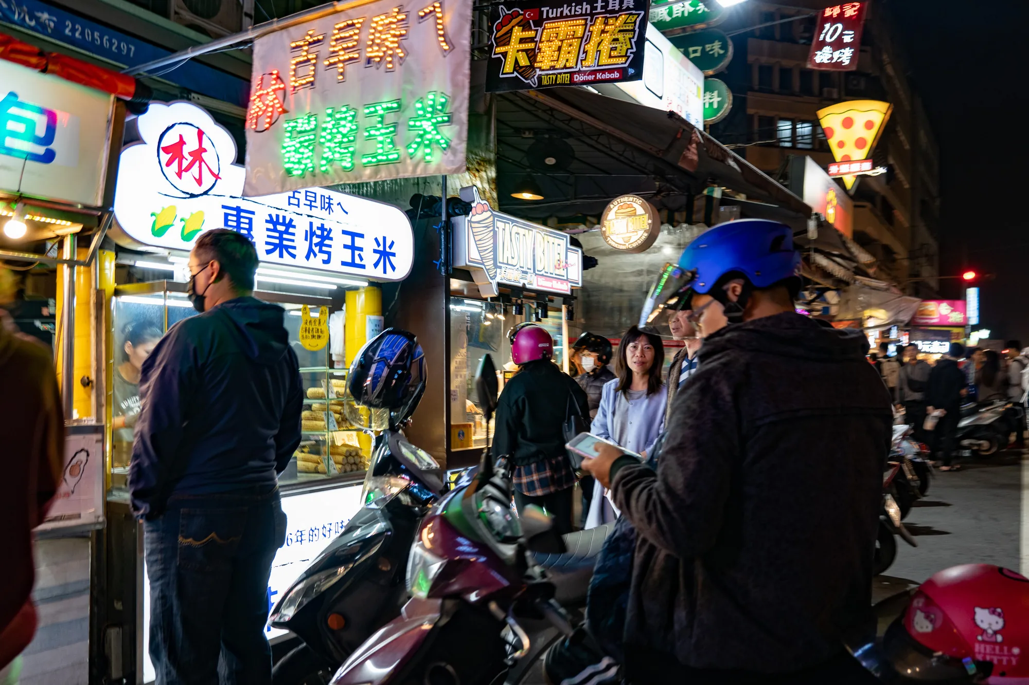 この写真は、台湾の台北で撮影された夜の屋台街の様子を捉えています。通りには様々な屋台が立ち並び、明るいネオンサインが点灯して活気にあふれています。写真の左側には「林 東業烤玉米」と書かれた看板の屋台があり、焼きトウモロコシを売っているようです。手前にはマスクをした男性が並んでおり、屋台で購入しようとしている様子が伺えます。他の屋台では「Turkish土耳其 Kebab」と書かれた看板があり、ケバブを売っていることがわかります。また、ピザの形をしたネオンサインも見え、様々な種類の食べ物が売られていることが分かります。道にはスクーターが停められており、多くの人々が行き交い、賑やかな雰囲気を醸し出しています。全体的に、台北の屋台街の活気と多様性を感じさせる一枚です。