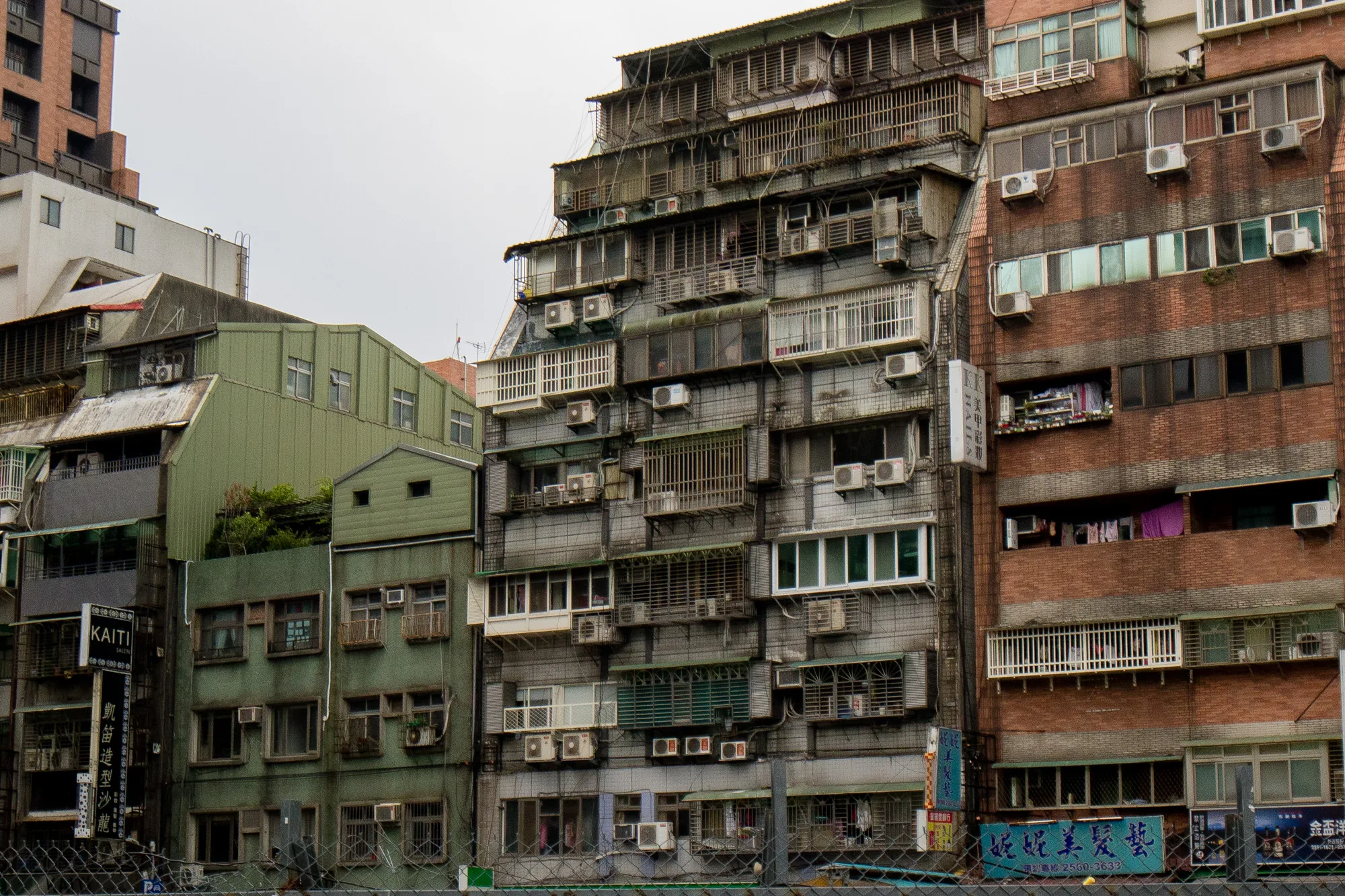 台湾の台北で撮影された写真です。画面全体に複数の建物が密集して写っており、空はどんよりとした曇り空です。建物はそれぞれ異なる色合いと構造をしています。左端には緑色の建物があり、その隣には灰色の建物がそびえ立っています。灰色の建物は、後から付け足されたような構造で、エアコンの室外機や鉄格子、ベランダなどが無秩序に取り付けられています。右端にはレンガ色の建物があり、こちらもエアコンの室外機が多数設置されています。建物の窓からは洗濯物が見え、生活感があります。手前のフェンス越しに撮影されており、画面下部にフェンスの一部が写っています。また、看板もいくつか確認でき、美容院のものなどが読み取れます。全体的に古びた印象で、台北の都市景観の一部を切り取ったような写真です。