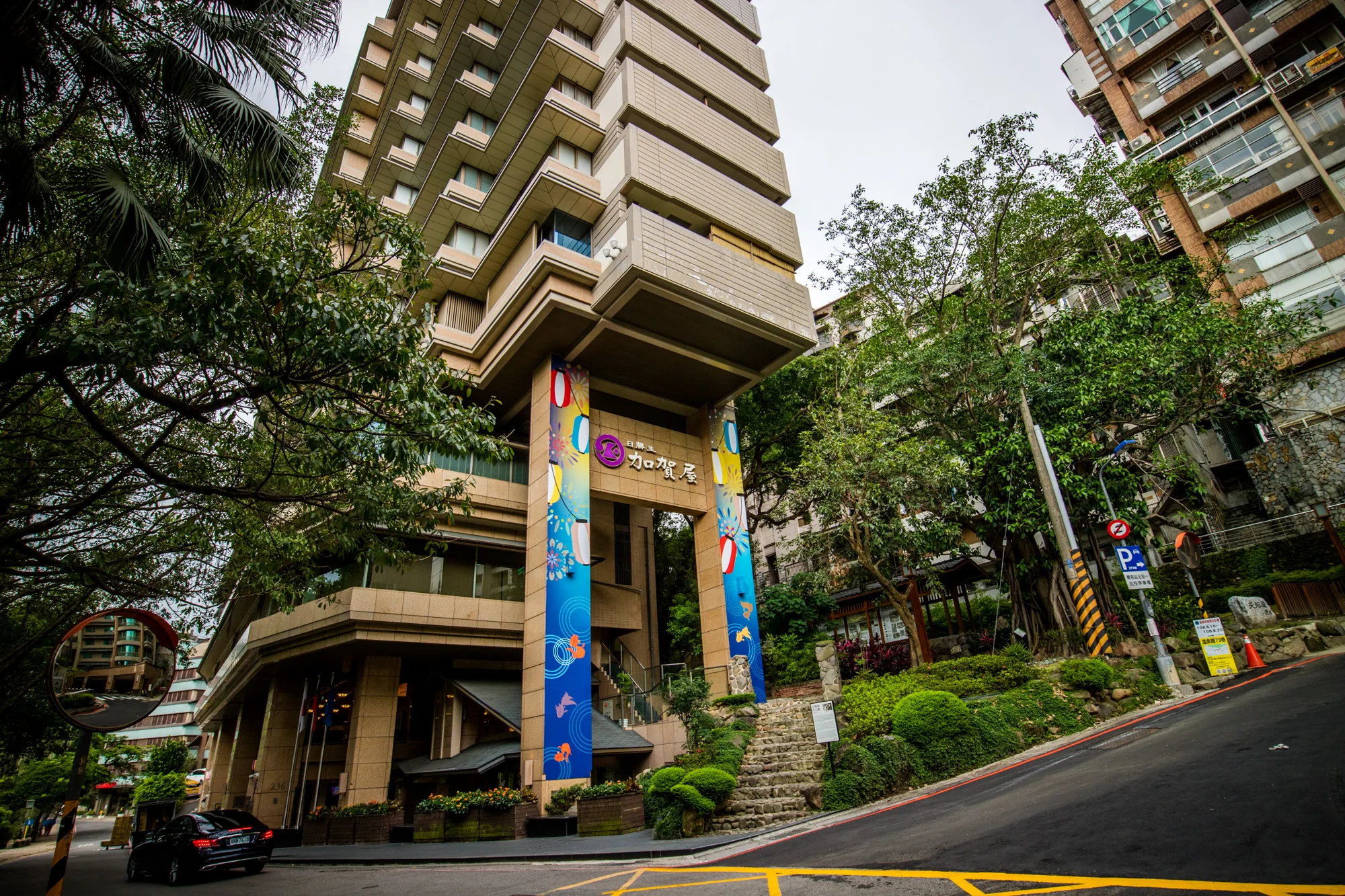 台湾の台北で撮影された写真です。主な被写体は「日勝生 加賀屋」という名前のホテルです。ホテルは特徴的な外観で、積み重ねられたような構造をしています。エントランスには、日本の提灯のようなデザインが施された青い垂れ幕が飾られています。エントランスへ続く階段があり、周りは緑豊かな植栽で彩られています。写真にはホテルに向かう坂道と、道路標識、一台の黒い車も写っています。全体的に落ち着いた雰囲気で、緑が多いことから、都会の中の静けさを感じさせる風景です。曇り空のため、全体的に少し暗めのトーンです。