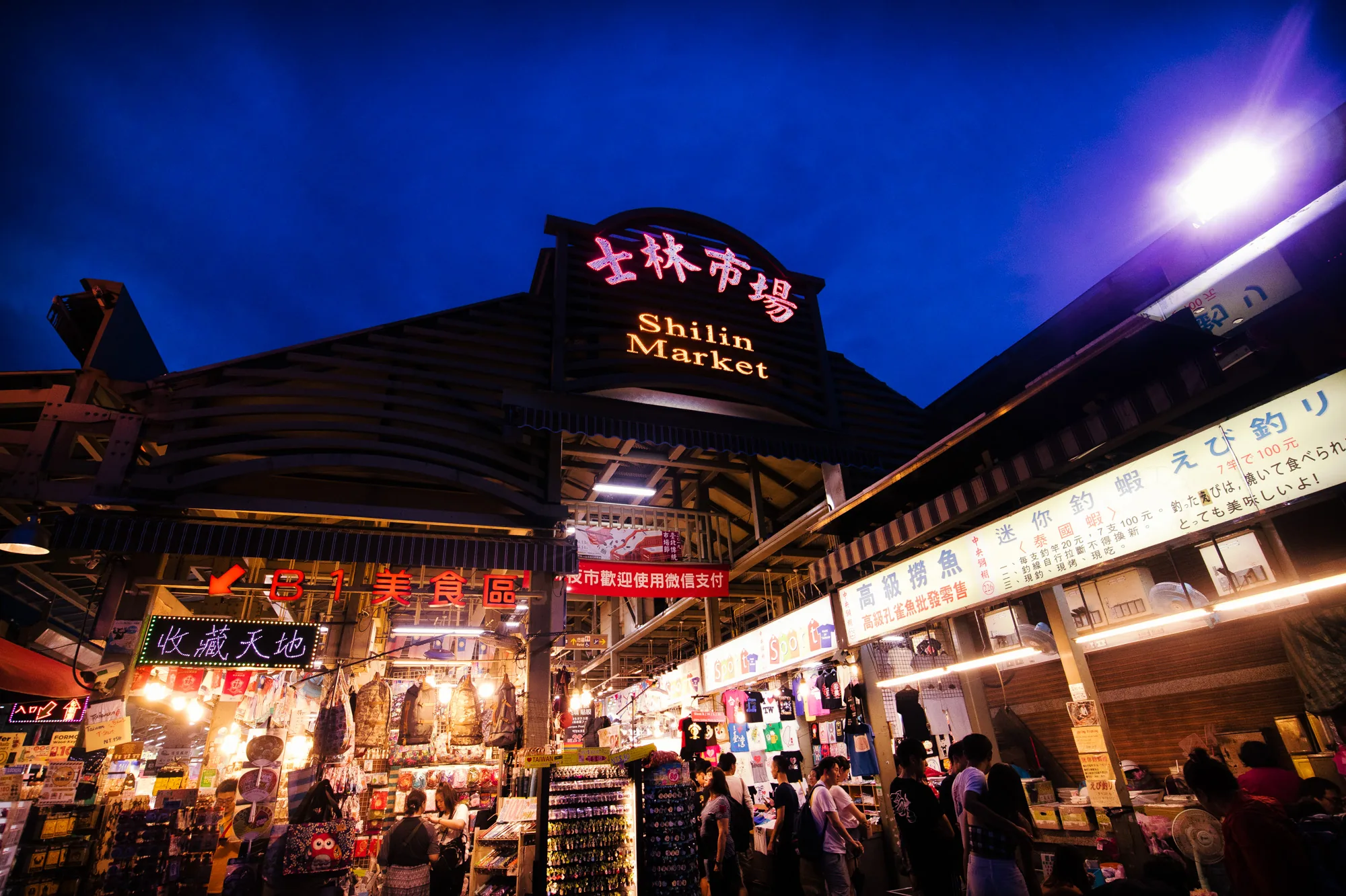 台湾の台北にある士林市場の夜の風景を捉えた写真です。空は濃い青色に染まり、市場の入り口を照らすライトが鮮やかに輝いています。「士林市場/Shilin Market」と書かれた看板が大きく掲げられており、その文字が赤とオレンジ色にライトアップされています。市場の内部は多くの人で賑わっており、様々な店舗が軒を連ねています。食べ物、衣類、お土産など、様々な商品が並んでいるのが見えます。看板やネオンサインが光り、活気ある市場の雰囲気を伝えています。