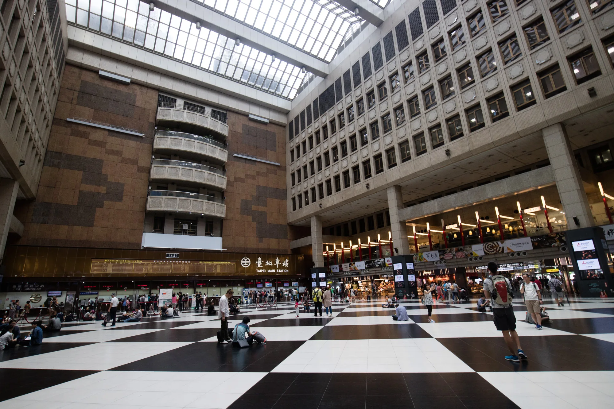 写真は、台湾の台北にある台北駅の内部を撮影したものです。天井が高く、ガラス張りの屋根から自然光が差し込む吹き抜け空間が印象的です。床は白と黒の市松模様になっており、モダンな雰囲気です。写真には、駅の出発案内板、切符売り場、様々な店舗が見られ、多くの人々が座ったり、歩き回ったりしています。人々は旅行者や地元住民など様々で、それぞれの目的を持って駅を利用している様子が伺えます。全体的に活気があり、都会の駅ならではのエネルギーを感じさせる写真です。