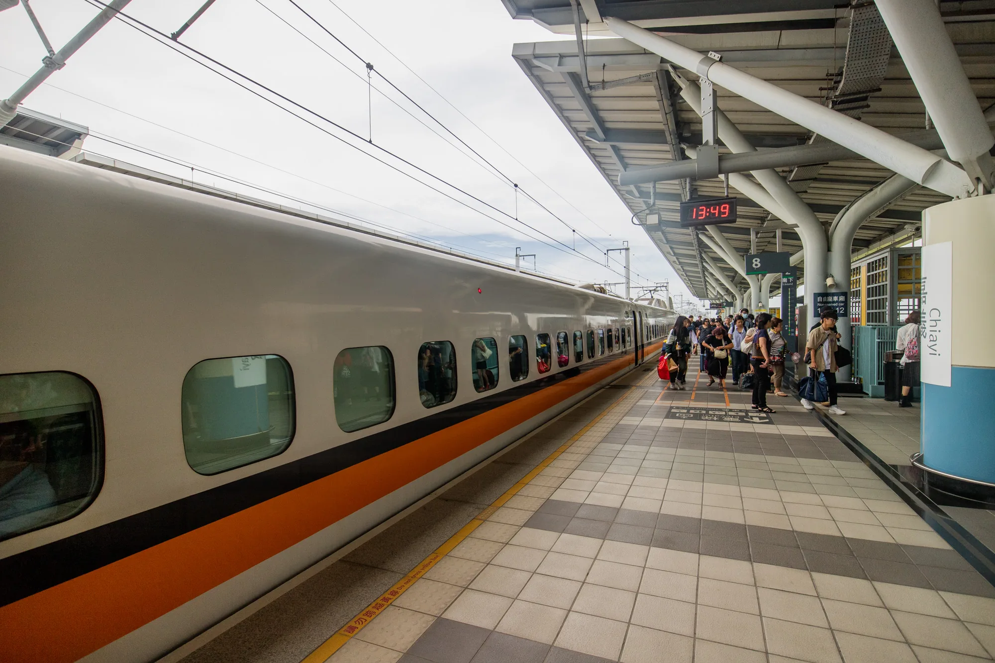 この写真は台湾の台北にある駅のホームの様子を捉えています。ホームには停車中の列車が写っており、その車体は白を基調とし、オレンジ色のラインが特徴的です。列車の窓からは乗客の姿がうかがえます。ホームには多くの人が列車を待っている様子で、様々な服装の人々が立っています。頭上には駅の屋根が広がり、時刻を示すデジタル表示板が13時49分を示しています。ホームの床はタイル張りで、黄色い線が引かれており、安全のための注意書きが見られます。駅のプラットフォーム番号は8番と表示されています。全体的に、台湾の鉄道の日常的な風景が写し出されています。