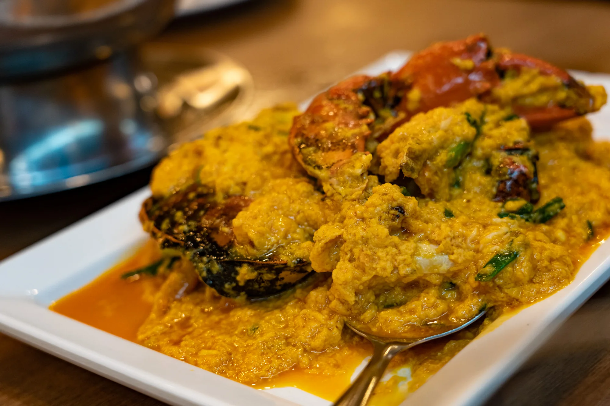 タイのバンコクで撮影された写真です。白い皿に盛られたプーパッポンカリー（蟹のカレー炒め）がメインで写っています。蟹は殻付きで、濃厚な黄色のカレーソースに覆われています。蟹の爪の部分が皿の上に突き出ており、食欲をそそります。カレーソースは、卵や香味野菜などが使われているようで、とろみがあります。皿の脇にはカレーソースが溜まっており、風味豊かな香りが伝わってくるようです。手前にはスプーンが添えられています。背景はぼけていますが、テーブルの木目と奥に他の食器類があることがわかります。全体的に暖色系の色調で、美味しそうな雰囲気が漂っています。