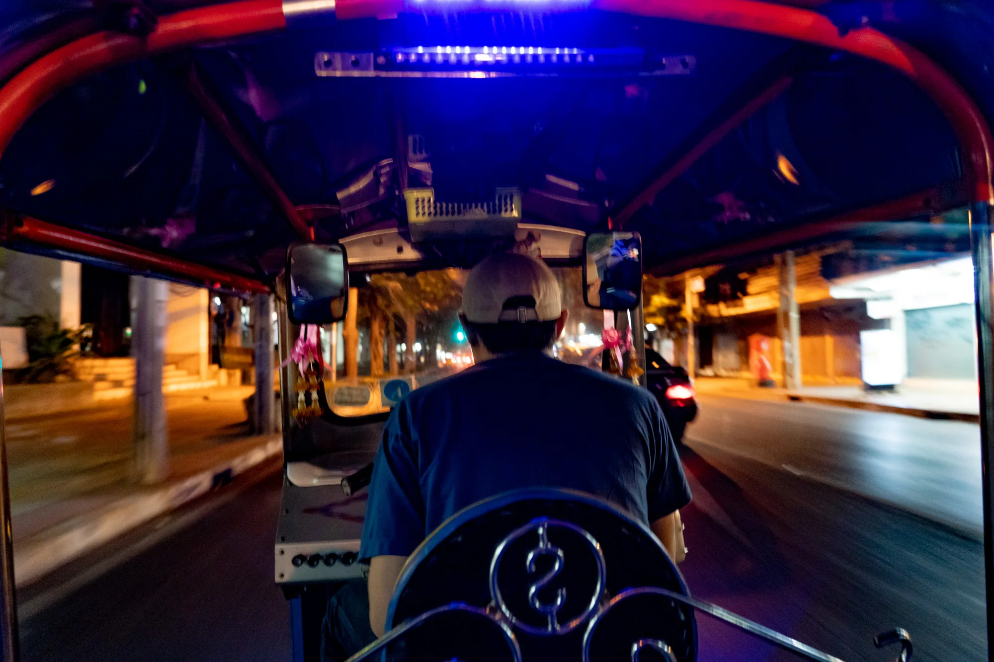 この写真は、タイのバンコクで撮影された、夜のトゥクトゥクからの眺めを捉えています。視点はトゥクトゥクの後部座席から運転手を見ている形で、運転手の背中、帽子、そして特徴的なハンドルが前景にあります。運転手は青いシャツを着て、ベージュの帽子を被っています。  トゥクトゥクの天井は赤と黒で装飾されており、青いLEDライトが光っています。サイドミラーには可愛らしい装飾が施されています。外の景色は夜の街並みで、オレンジ色の街灯が道路を照らしています。動いているため、建物や街路樹はぼやけて見えます。車のヘッドライトやテールライトもちらほら見え、活気のある夜のバンコクの雰囲気を醸し出しています。写真全体は少しぶれており、トゥクトゥクの疾走感を表しています。