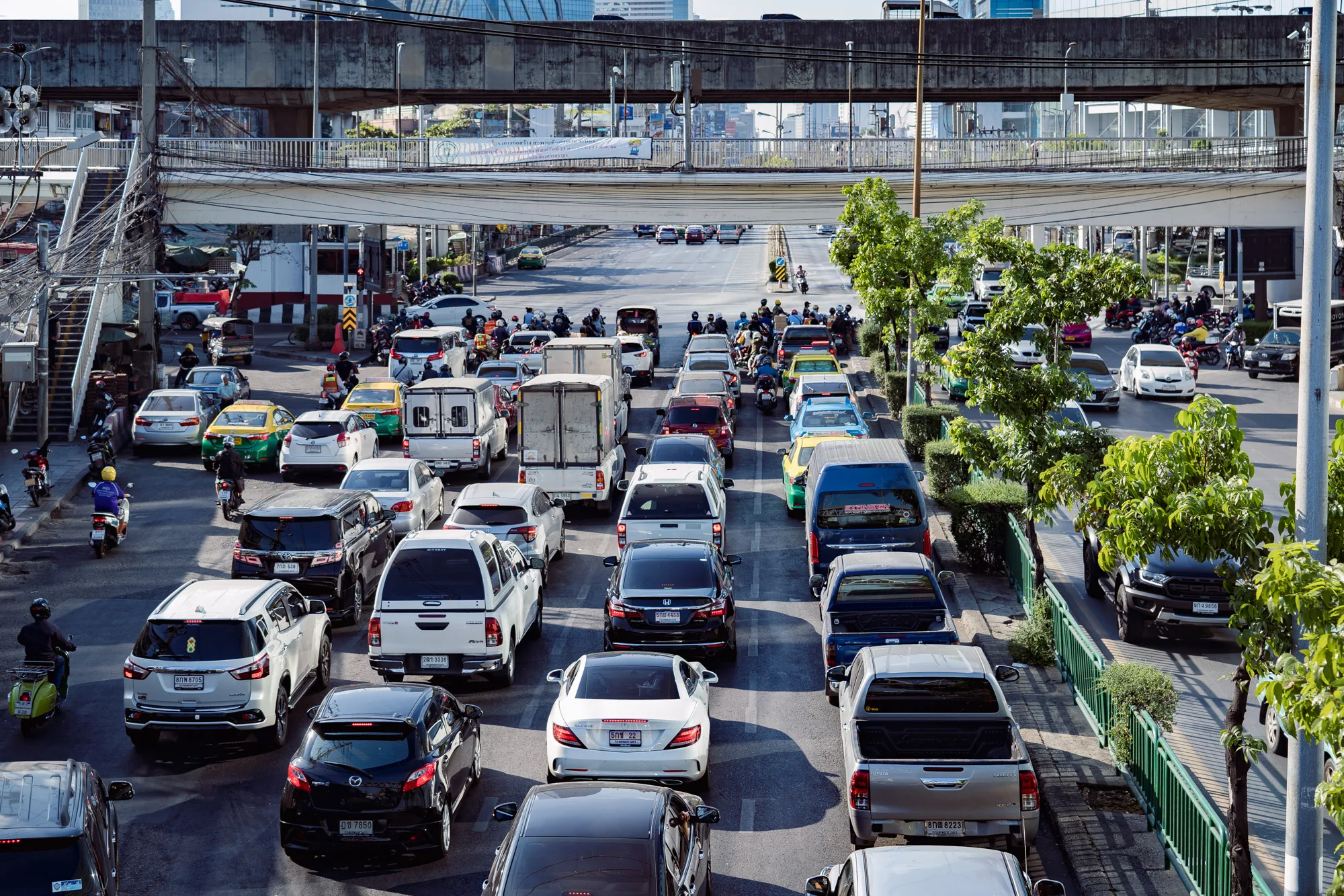 写真はタイのバンコクで撮影された、交通渋滞の様子を捉えたものです。複数の車線には車、トラック、タクシー、バイクが密集して走行しており、特に手前の車線は渋滞でほとんど動いていないように見えます。タクシーは特徴的な黄緑色で、バンコクらしい風景を演出しています。歩道には歩道橋の階段が設置されており、道路を横断する人が利用できるようになっています。高架道路も写っており、その上にも車両が走行しているのが見えます。背景には高層ビル群がそびえ立ち、バンコクの都市景観を象徴しています。全体的に明るい日中の光が差し込み、交通量の多さと合わせて活気のある雰囲気を感じさせます。